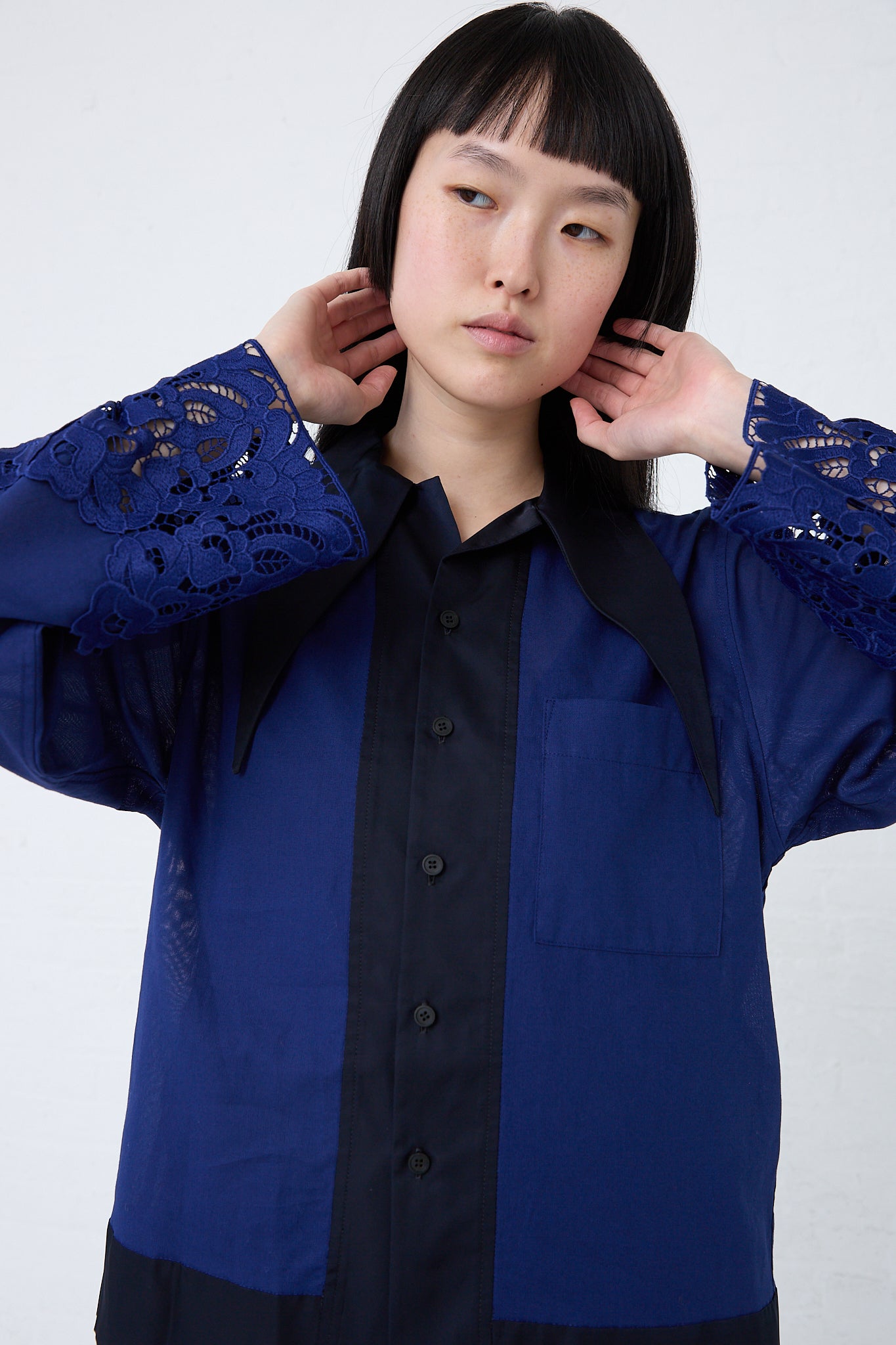 Woman in a TOGA ARCHIVES Mesh Lace Shirt in Blue with relaxed fit, touching her ears, against a white background.