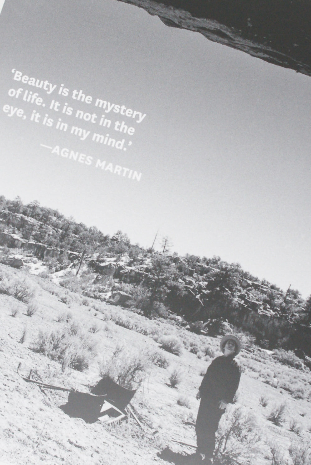 A monochrome image of a person standing in a desert landscape, with a quote by Agnes Martin about beauty being a mystery of life, captured during a retrospective exhibition from the Agnes Martin: Illustrated Monograph by Artbook/D.A.P.