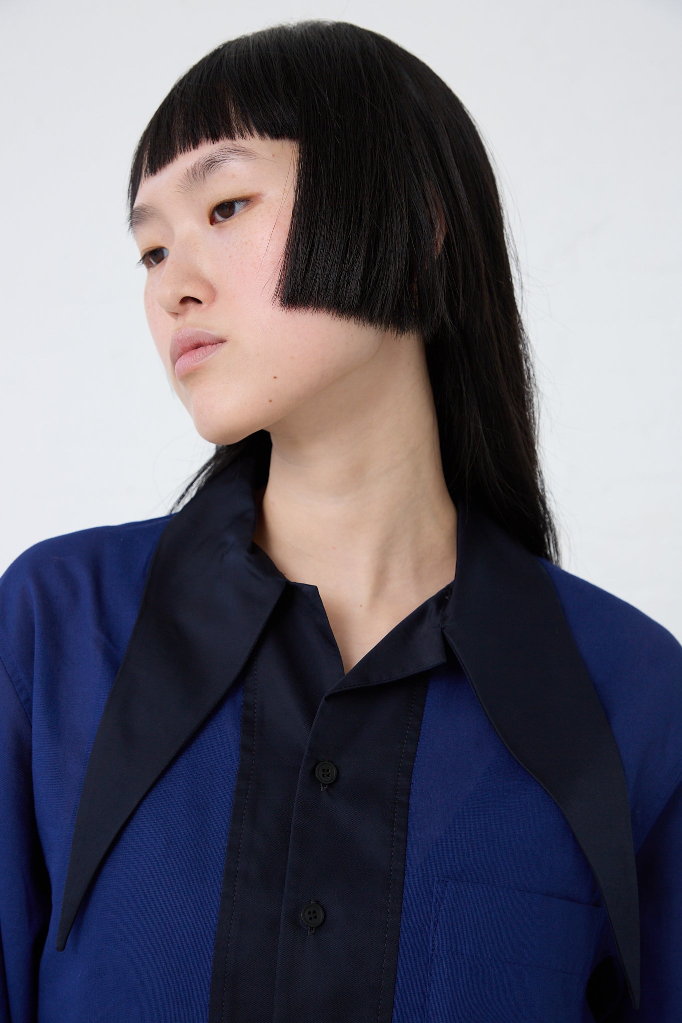 A young Asian woman with a blunt bob haircut, wearing a TOGA ARCHIVES Mesh Lace Shirt in Blue with a black collar, gazes to the side against a white background.