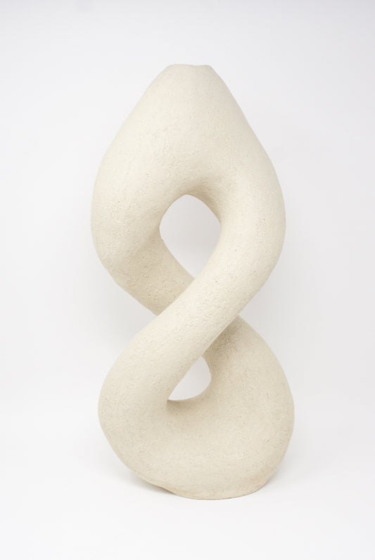 A beige, abstract sculptural form resembling a twisted loop with a rough texture against a plain white background, evocative of floral arrangements, titled "Hand Built Vessel No. 000395" by Lost Quarry.