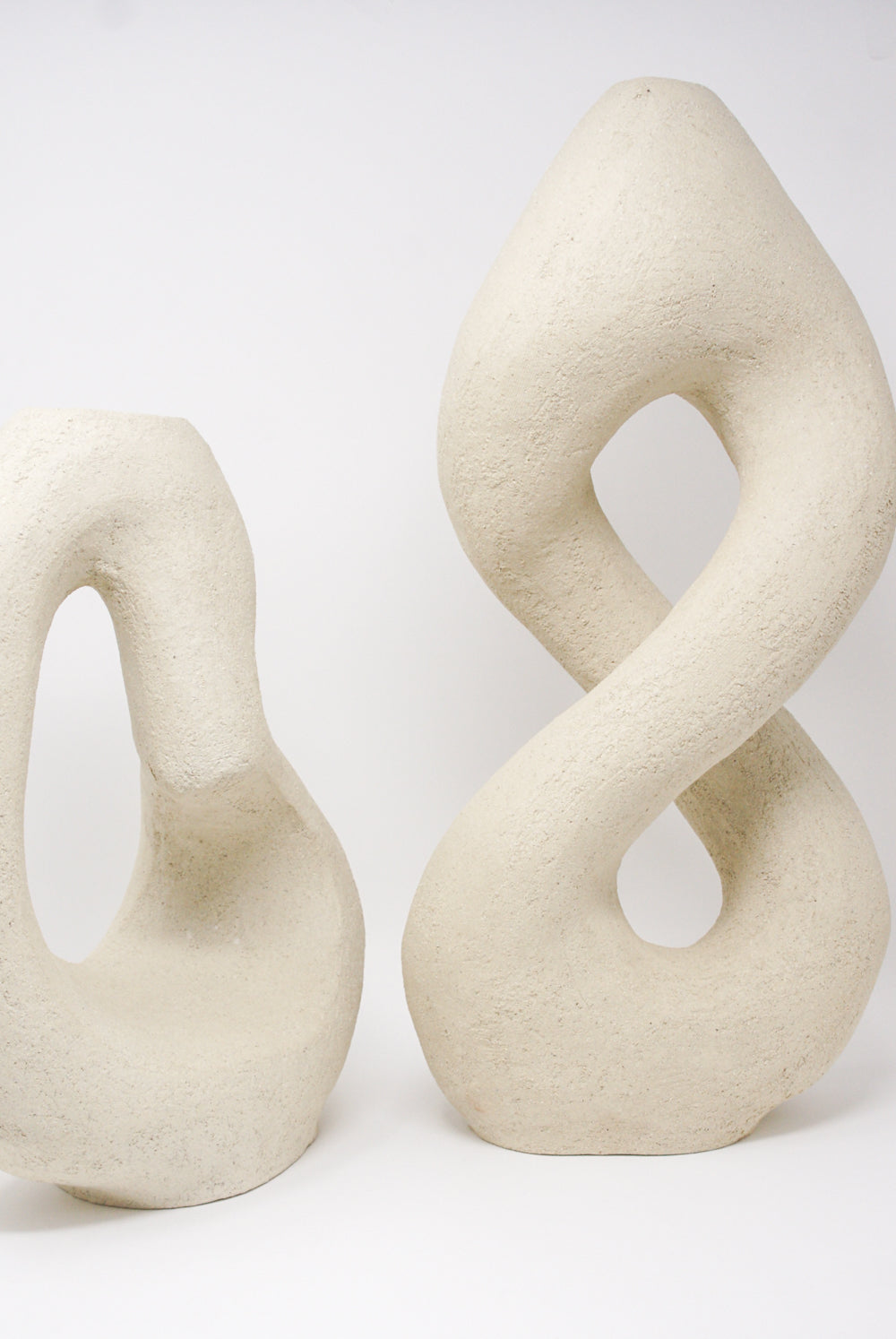 Two Hand Built Vessel No. 000395 sculptures by Lost Quarry, displaying curved, looped forms in an abstract beige design against a simple white background, evoke the elegance of artisan craftsmanship.
