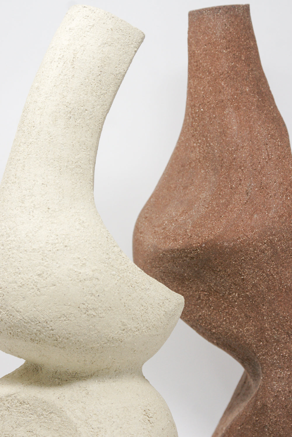 Two abstract, curvy sculptures stand against a plain background. The left sculpture is beige with a rough texture, similar to the handcrafted design of Lost Quarry's Hand Built Vessel No. 000345, while the right sculpture is brown and also features a comparable texture.