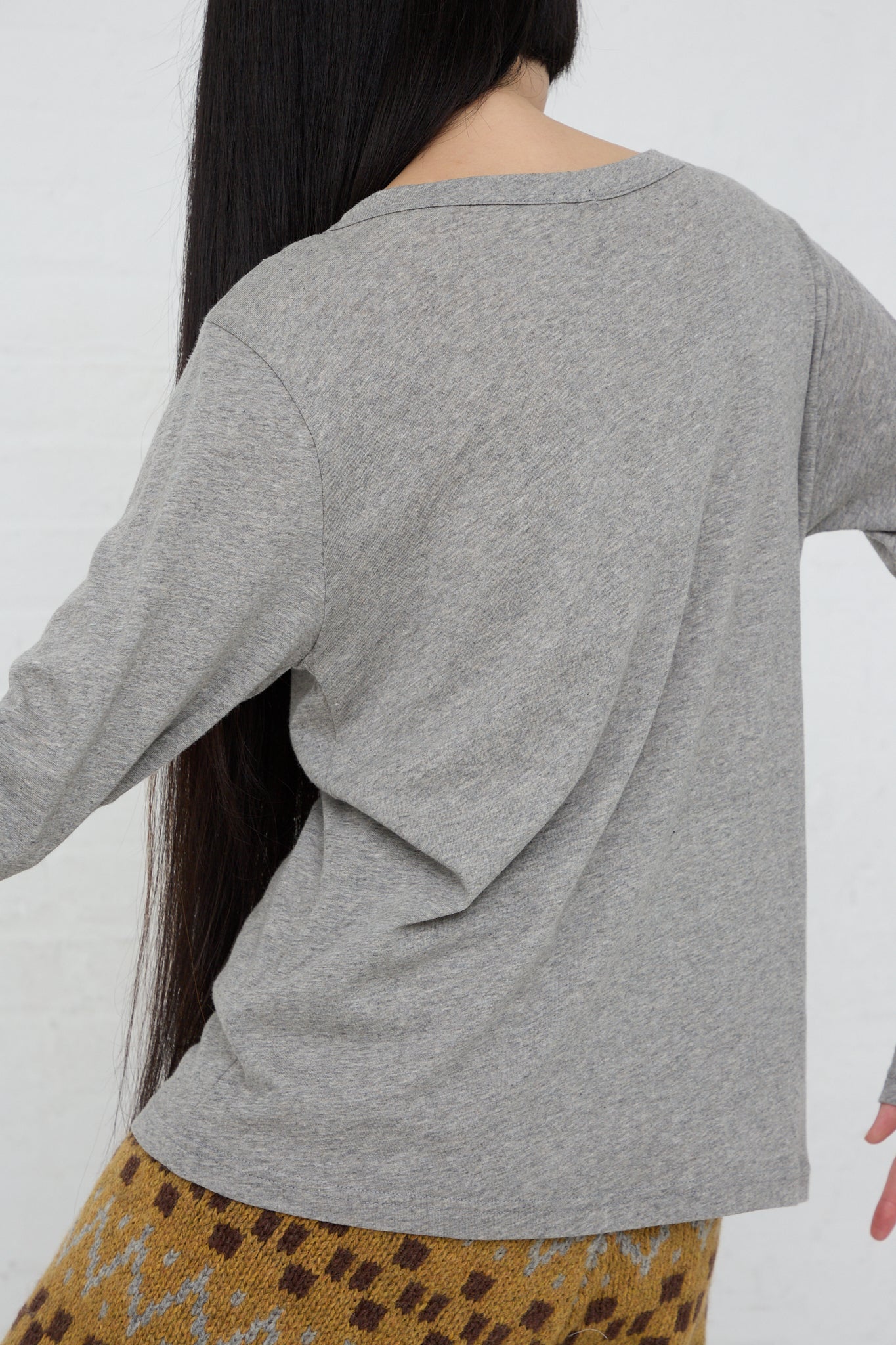 A woman wearing a gray long-sleeved Cotton Knit Pullover by Ichi and a patterned skirt, styled in a relaxed fit. Back view.