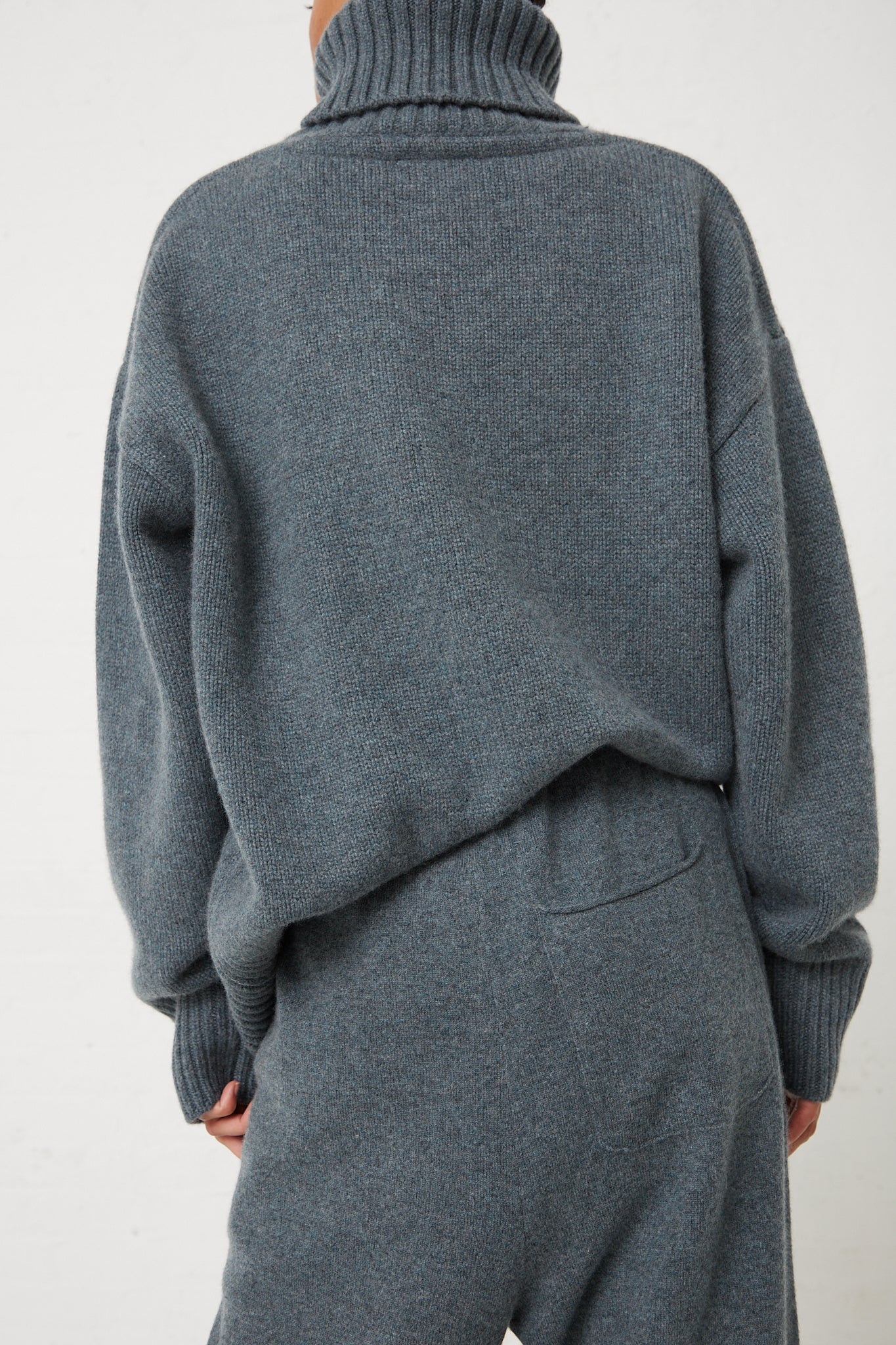 The back view of a woman wearing a grey turtleneck sweater and Extreme Cashmere's No. 320 Rush Trouser in Wave with drawstring waist.