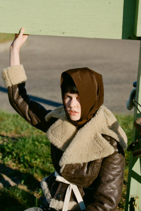 A woman with a leather and fur jacket on with her hand in the air holding onto a structure. Click here to read more.