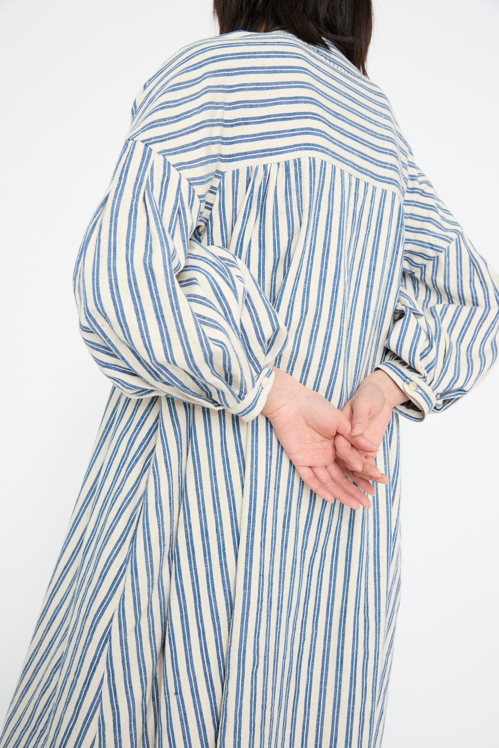 Someone in the 11.11 Cotton Gurue Dress, Medium Indigo Stripe, with an elegant A-line silhouette, is viewed from the back with hands clasped behind.