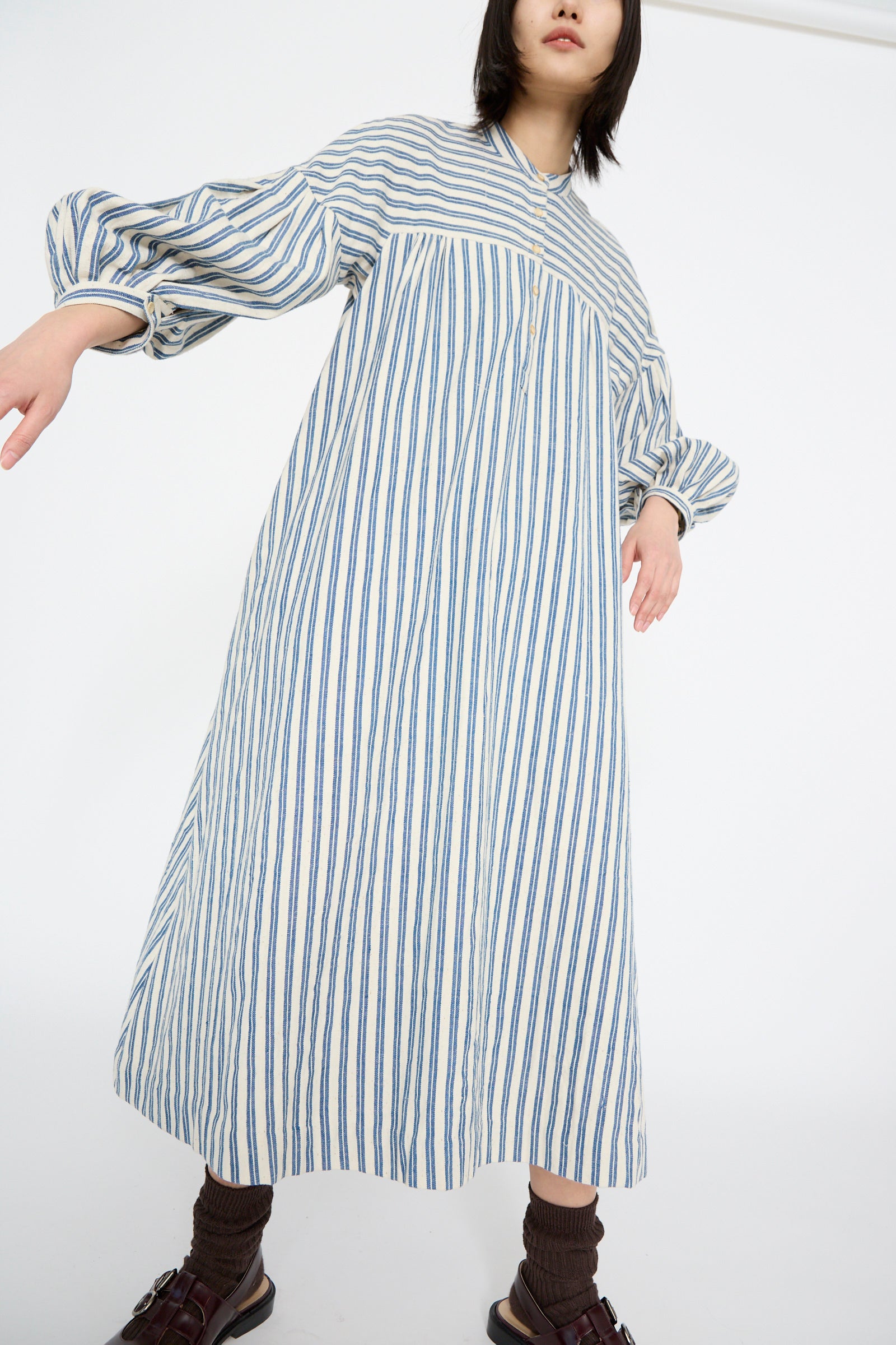 Wearing the 11.11 Cotton Gurue Dress in Medium Indigo Stripe, a person elegantly pairs it with brown socks and sandals. They stand with one arm slightly extended against a plain background. Made from handwoven cotton, this dress features a long, loose-fitting A-line silhouette.