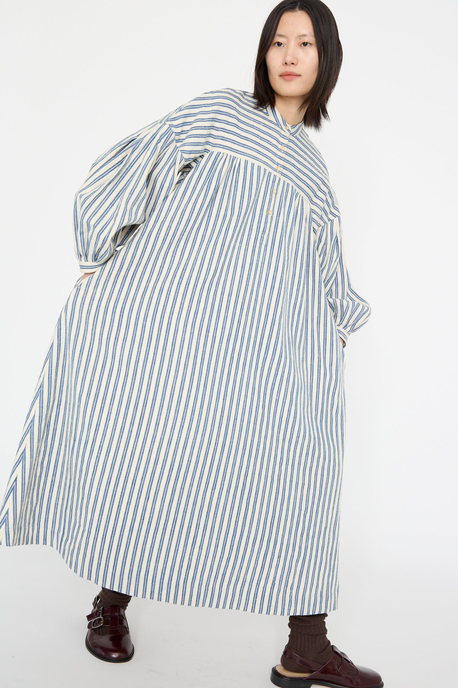 A person wearing the 11.11 Cotton Gurue Dress in Medium Indigo Stripe with an A-line silhouette and brown sandals against a plain background.