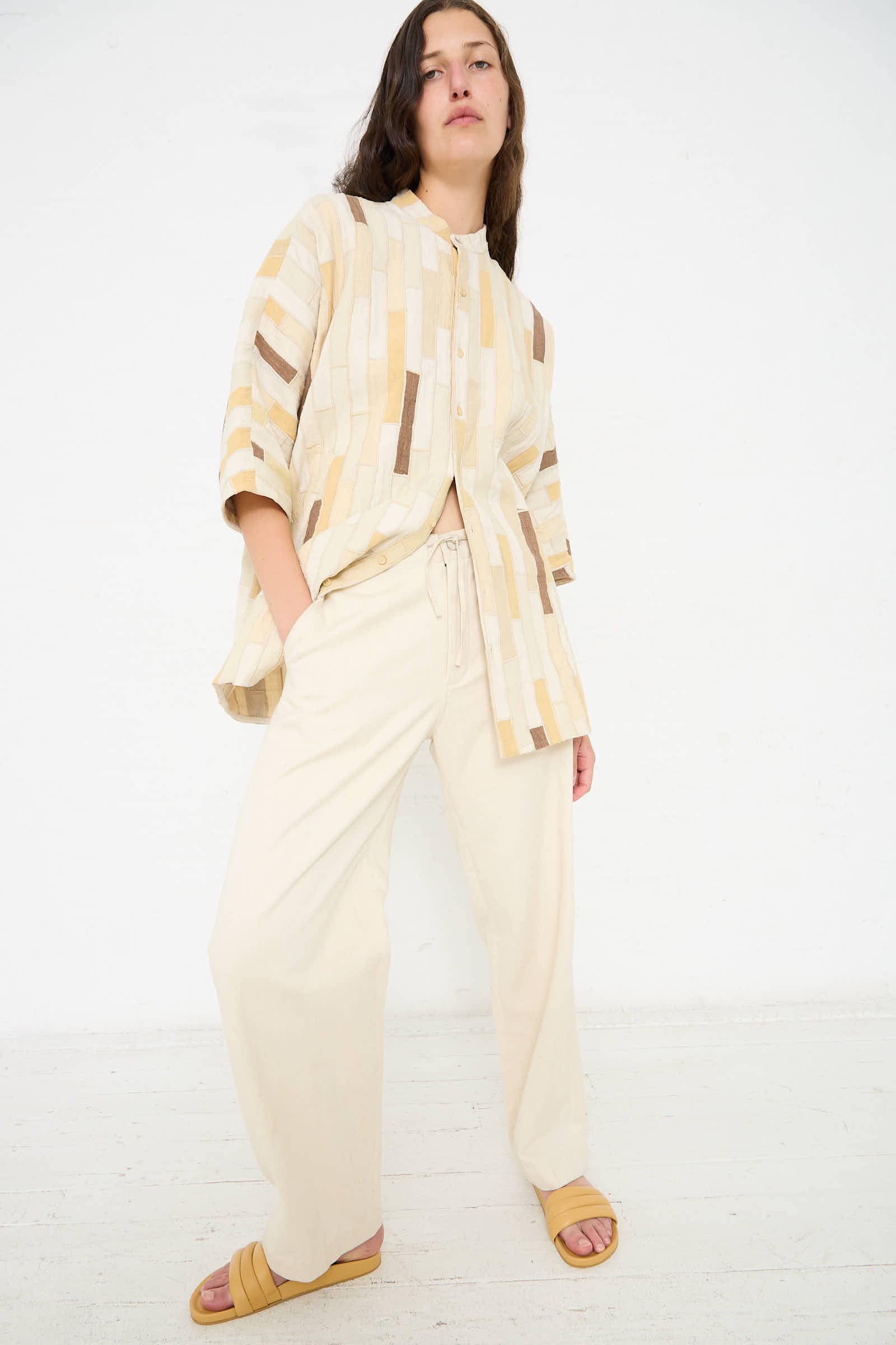 Person standing against a white background, wearing a beige patterned top, 11.11 Cotton Pant in Ecru with a relaxed fit, and open-toe sandals.