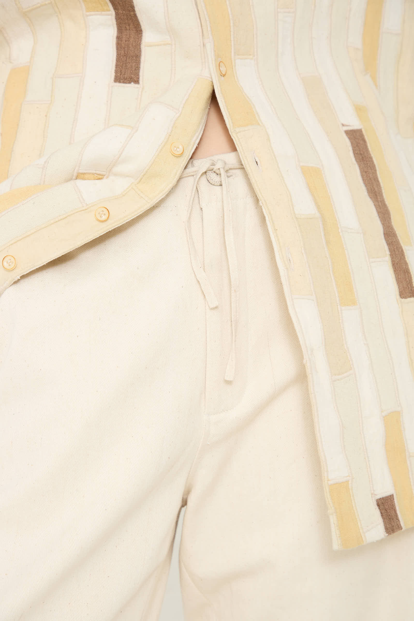 Close-up of a person wearing relaxed fit, light-colored *11.11 Cotton Pant in Ecru* with a drawstring and a buttoned, patterned shirt. The focus is on the waist area, showcasing the pull-on style pant with the shirt partially unbuttoned.