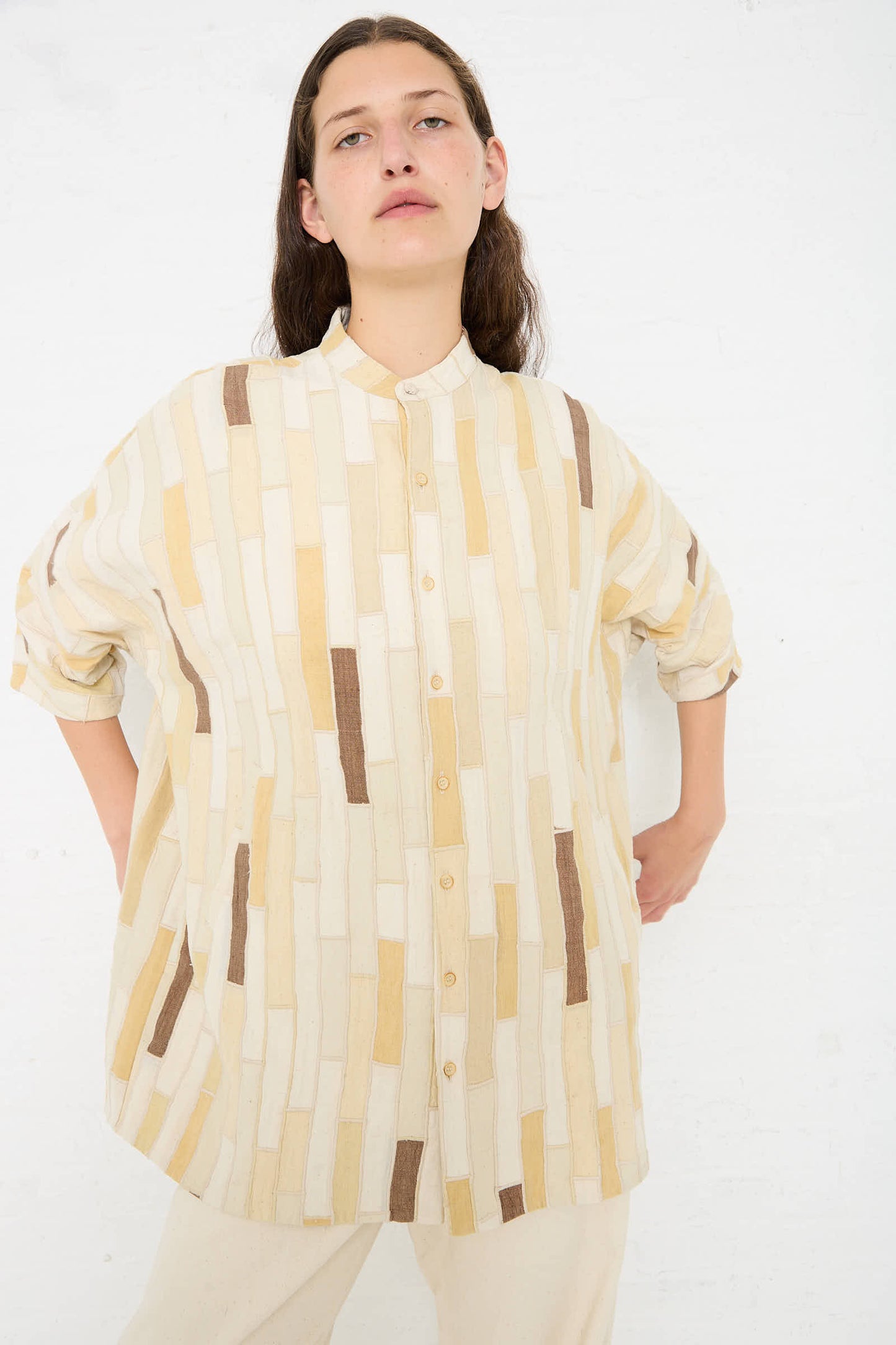 A person with long dark hair wears a loose, button-up Cotton Patchwork Statement Shirt in Shades of Ecru by 11.11 with a geometric pattern of beige, brown, and yellow and relaxed fit dolman sleeves, standing against a plain white background.