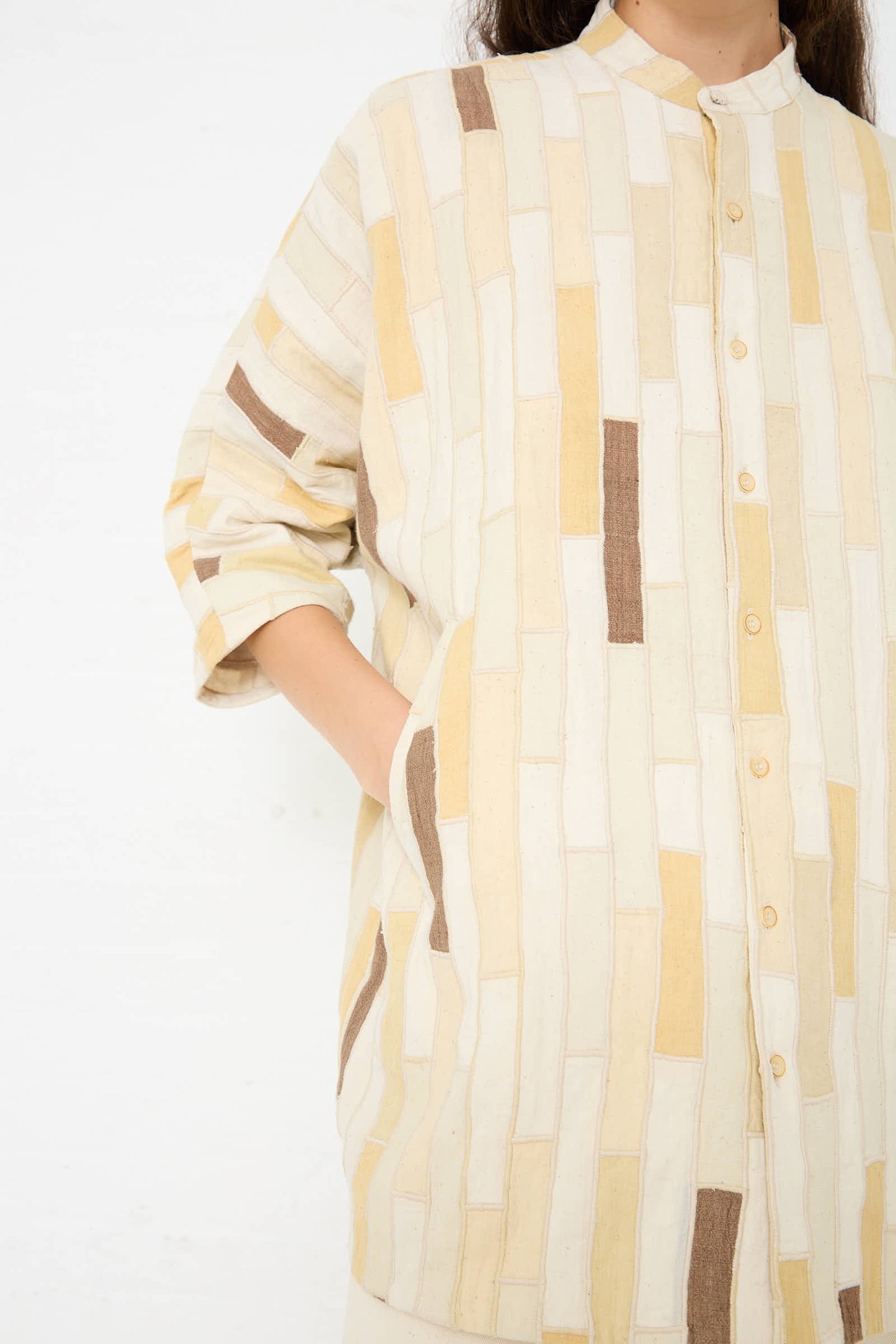 Person wearing a long-sleeved, button-up 11.11 Cotton Patchwork Statement Shirt in Shades of Ecru with a geometric pattern of beige, cream, and brown rectangles. The textured patchwork cotton top has a relaxed fit, and their left hand is in the shirt pocket. The background is a plain white wall.