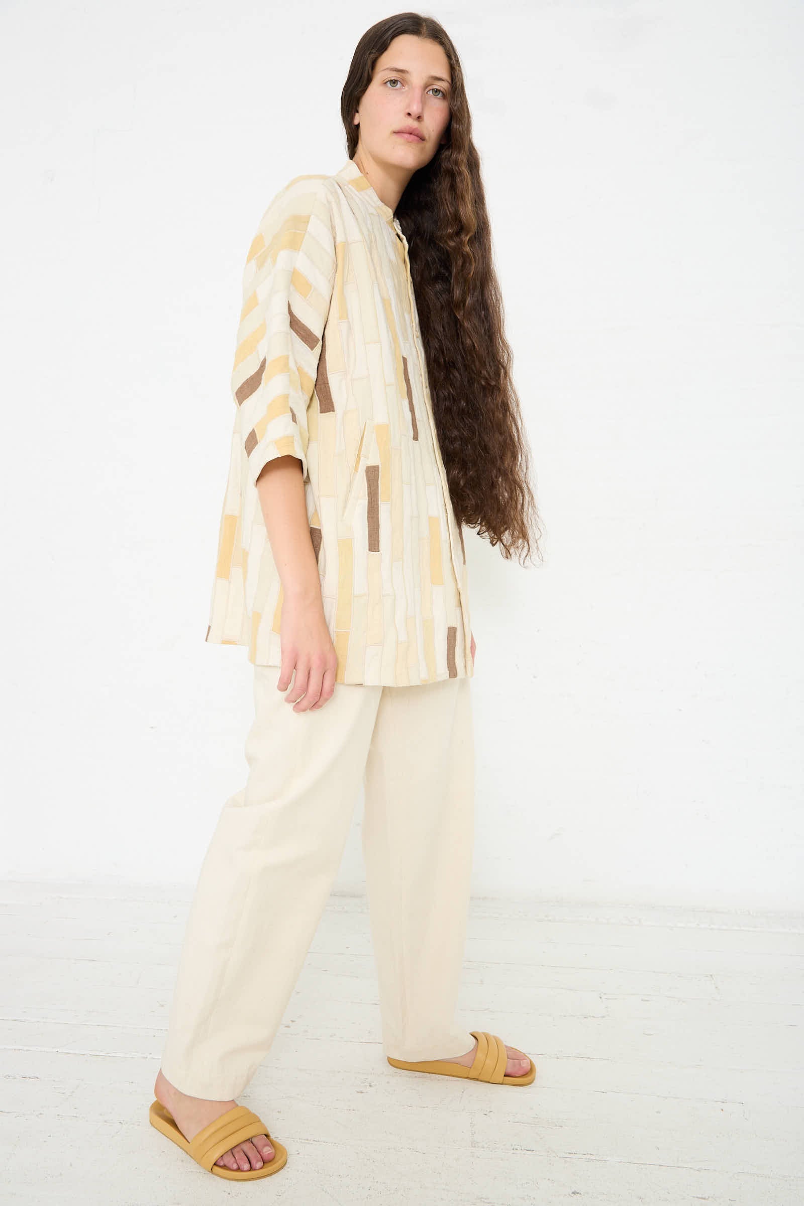 A person with long hair stands against a white background wearing a beige, Cotton Patchwork Statement Shirt in Shades of Ecru by 11.11 with dolman sleeves, light-colored pants, and yellow sandals.