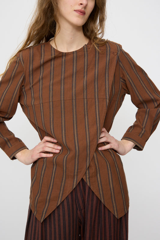 A woman poses with hands on hips, wearing the 11.11 Cotton Rubai Shirt in Filter Coffee—a relaxed fit, striped organic cotton long-sleeve shirt with an asymmetrical hem—paired with matching striped pants.