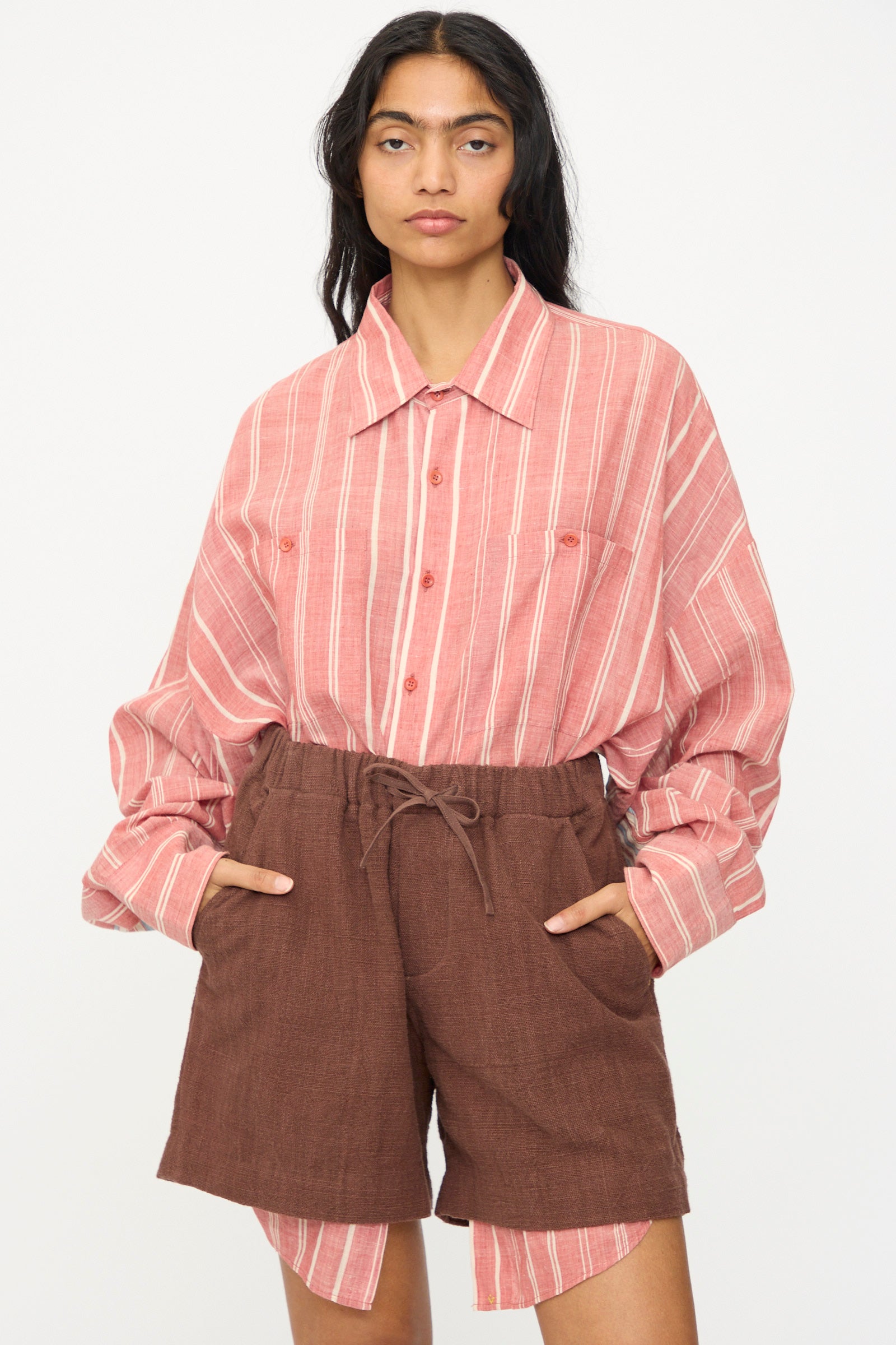 A person stands with hands in pockets, wearing the 11.11 Cotton Short in Dark Brown and a pink and white striped button-up shirt against a plain white background.
