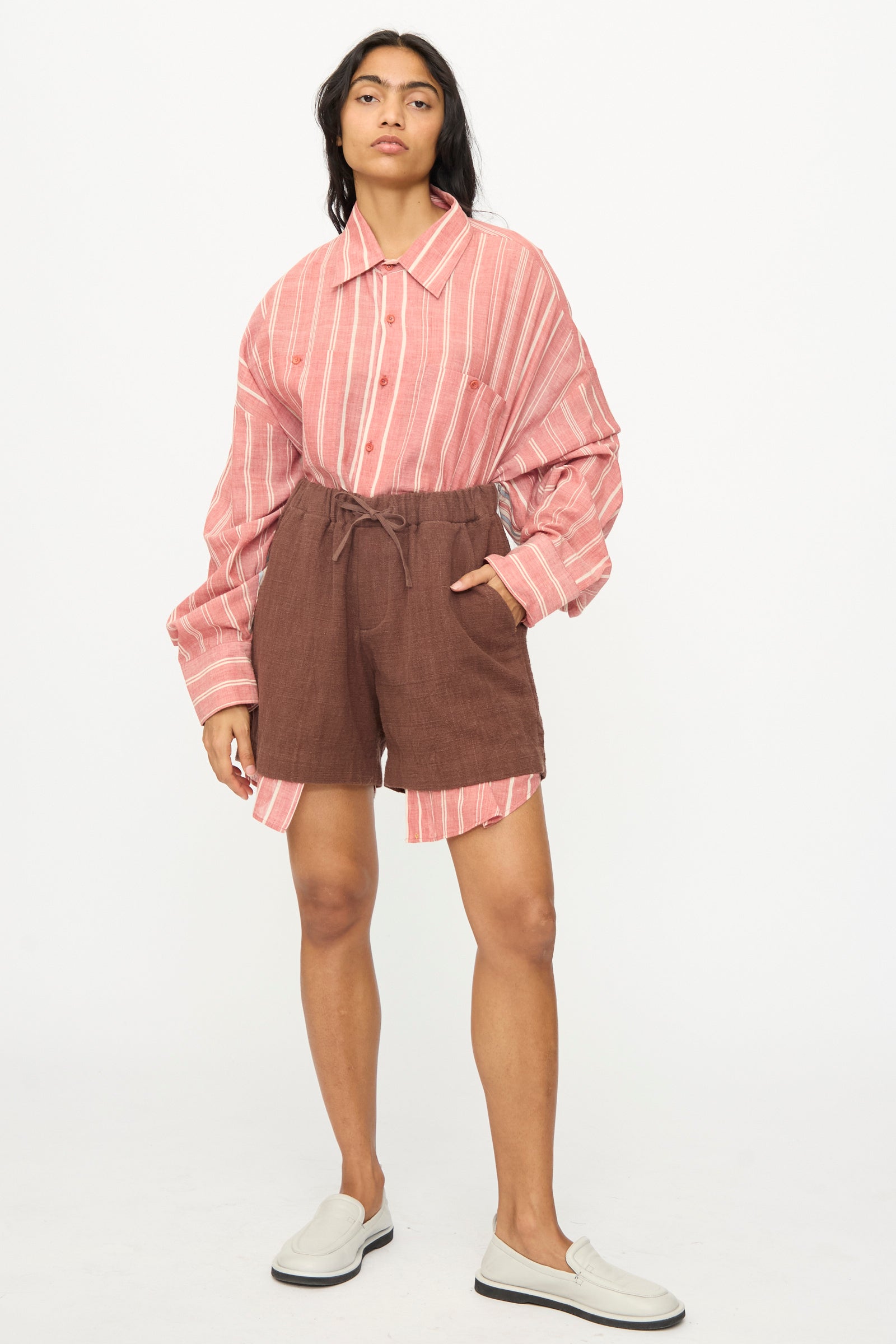 A person stands against a plain background wearing an oversized pink and white striped shirt, 11.11 Cotton Short in Dark Brown with a drawstring waist, and white slip-on shoes.