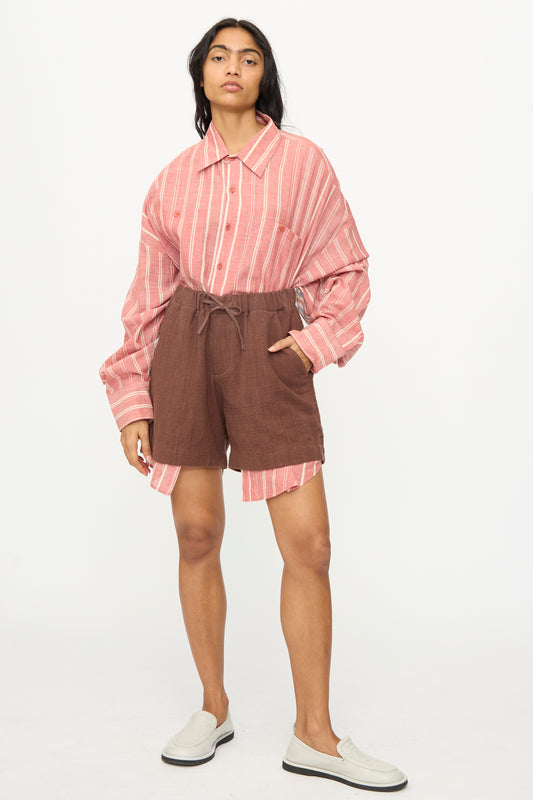 A person stands against a plain background wearing an oversized pink and white striped shirt, 11.11 Cotton Short in Dark Brown with a drawstring waist, and white slip-on shoes.
