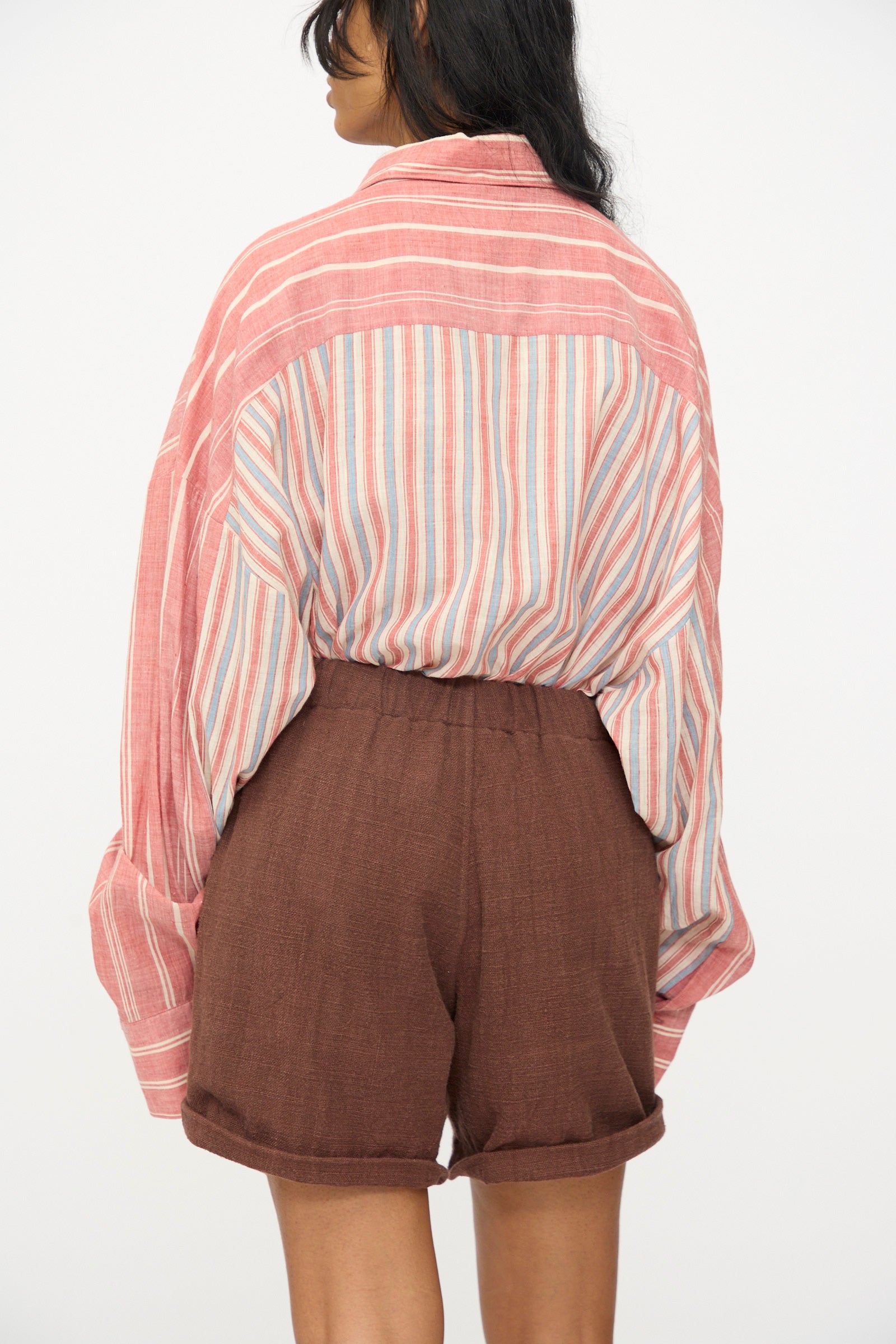 A person faces away, wearing 11.11’s Cotton Short in Dark Brown—high-waisted with a relaxed fit—paired with a loose, long-sleeved red and white striped shirt, set against a plain light background.