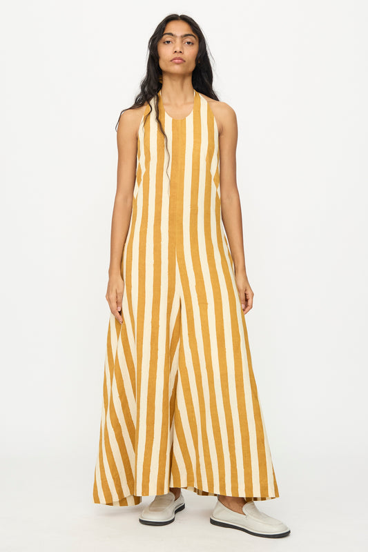 A person stands facing forward, wearing the 11.11 Cotton Stripe Jumpsuit in Mustard Yellow, featuring a sleeveless, wide-leg design in handwoven cotton with white stripes, paired with white slip-on shoes against a plain white background.
