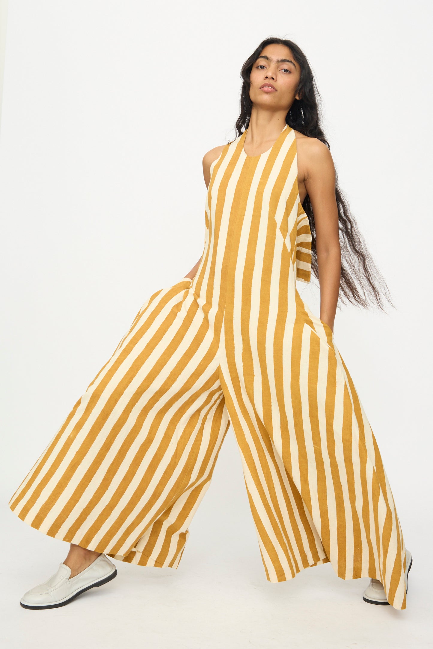 A woman models the 11.11 Cotton Stripe Jumpsuit in Mustard Yellow, featuring wide legs and a sleeveless handwoven cotton design with vertical stripes, posing with hands in pockets against a plain white background.