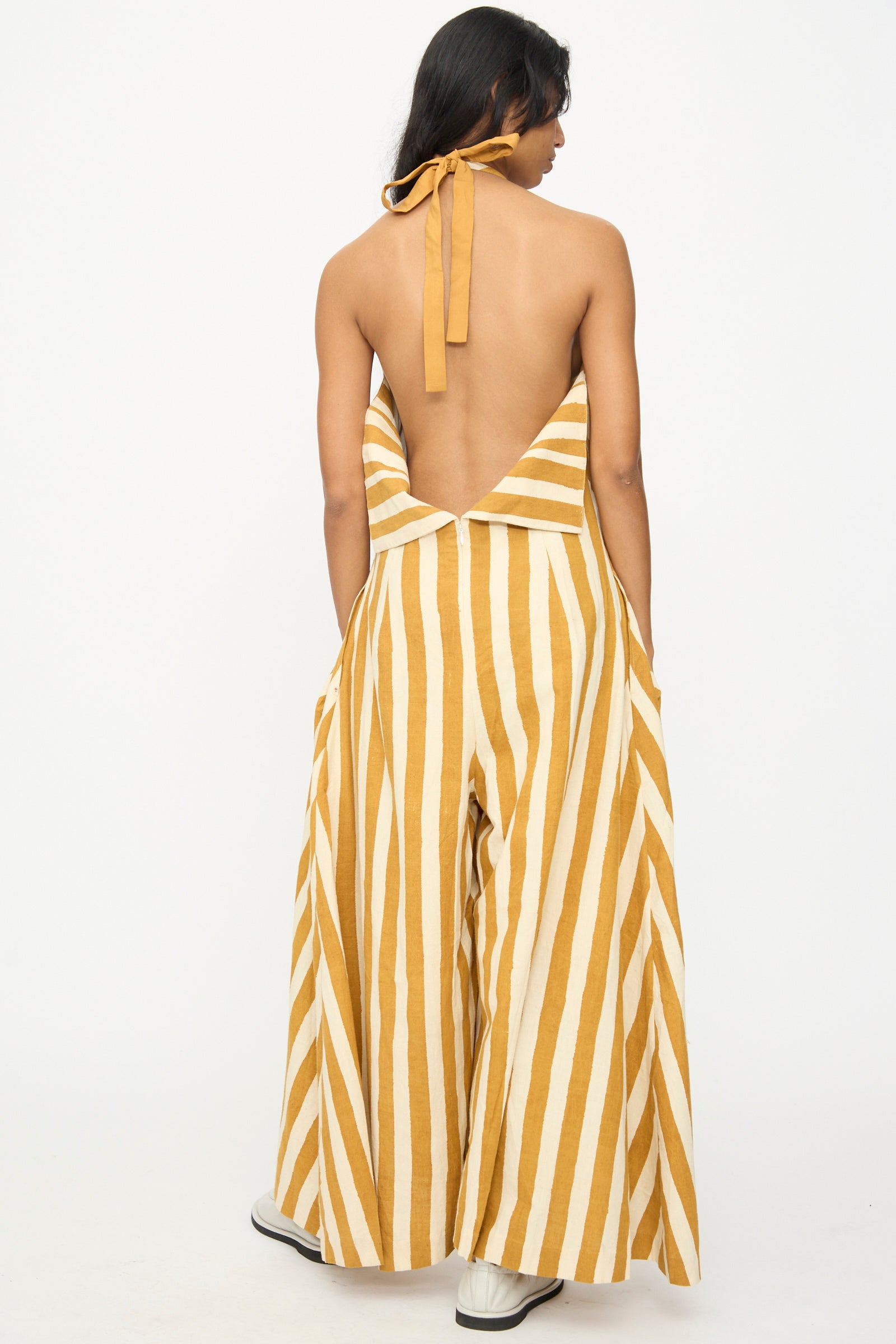 A person models the 11.11 Cotton Stripe Jumpsuit in Mustard Yellow, featuring wide mustard yellow and white vertical stripes and a backless halter-neck design, set against a plain white background.