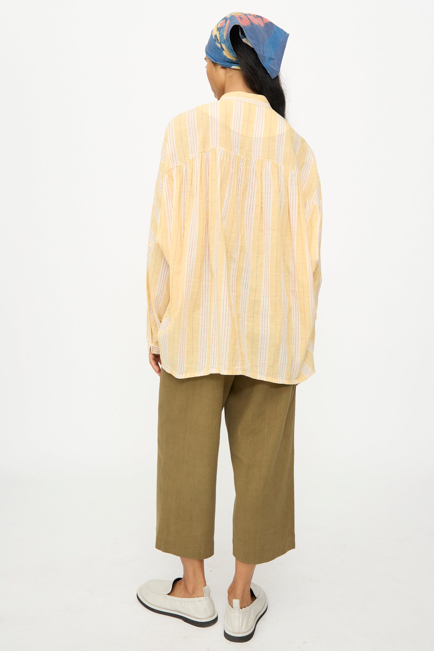 Person facing away in 11.11's Cotton Stripe Shirt in Mango Yellow, olive cropped pants, white slip-on shoes, and a blue patterned headscarf, against a plain white background.