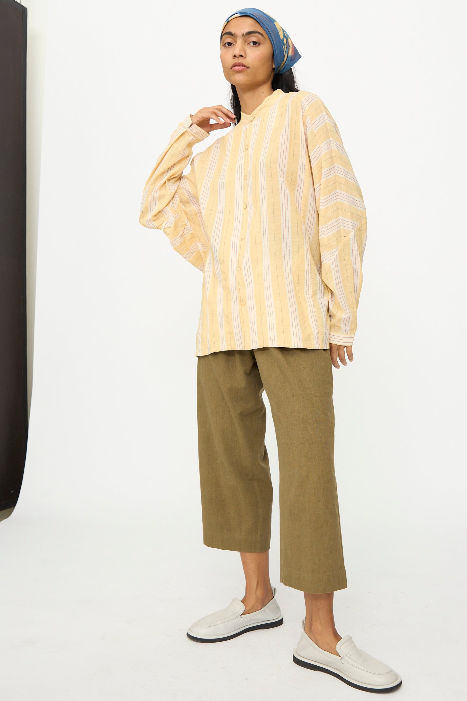 A person in the 11.11 Cotton Stripe Shirt in Mango Yellow, olive cropped pants, white slip-on shoes, and a blue headscarf stands against a plain white background.