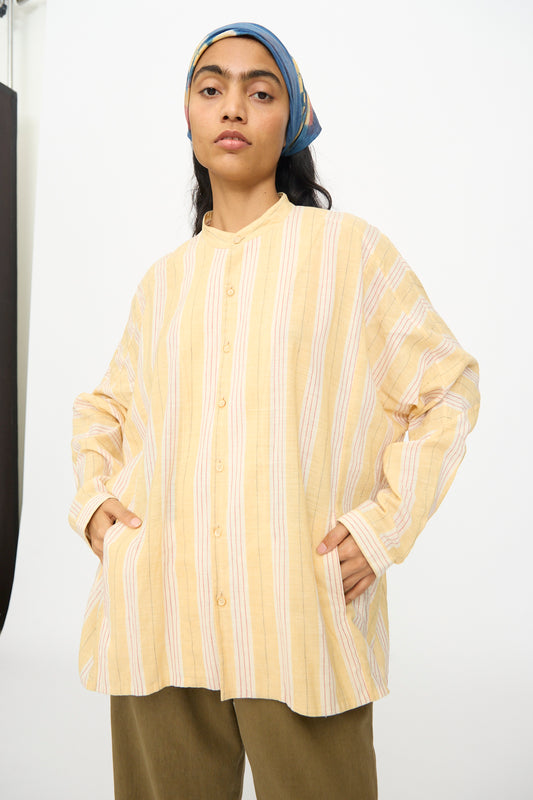A person wears the 11.11 Cotton Stripe Shirt in Mango Yellow, featuring a band collar, with olive pants and a blue headscarf, standing hands-in-pockets against a plain background.