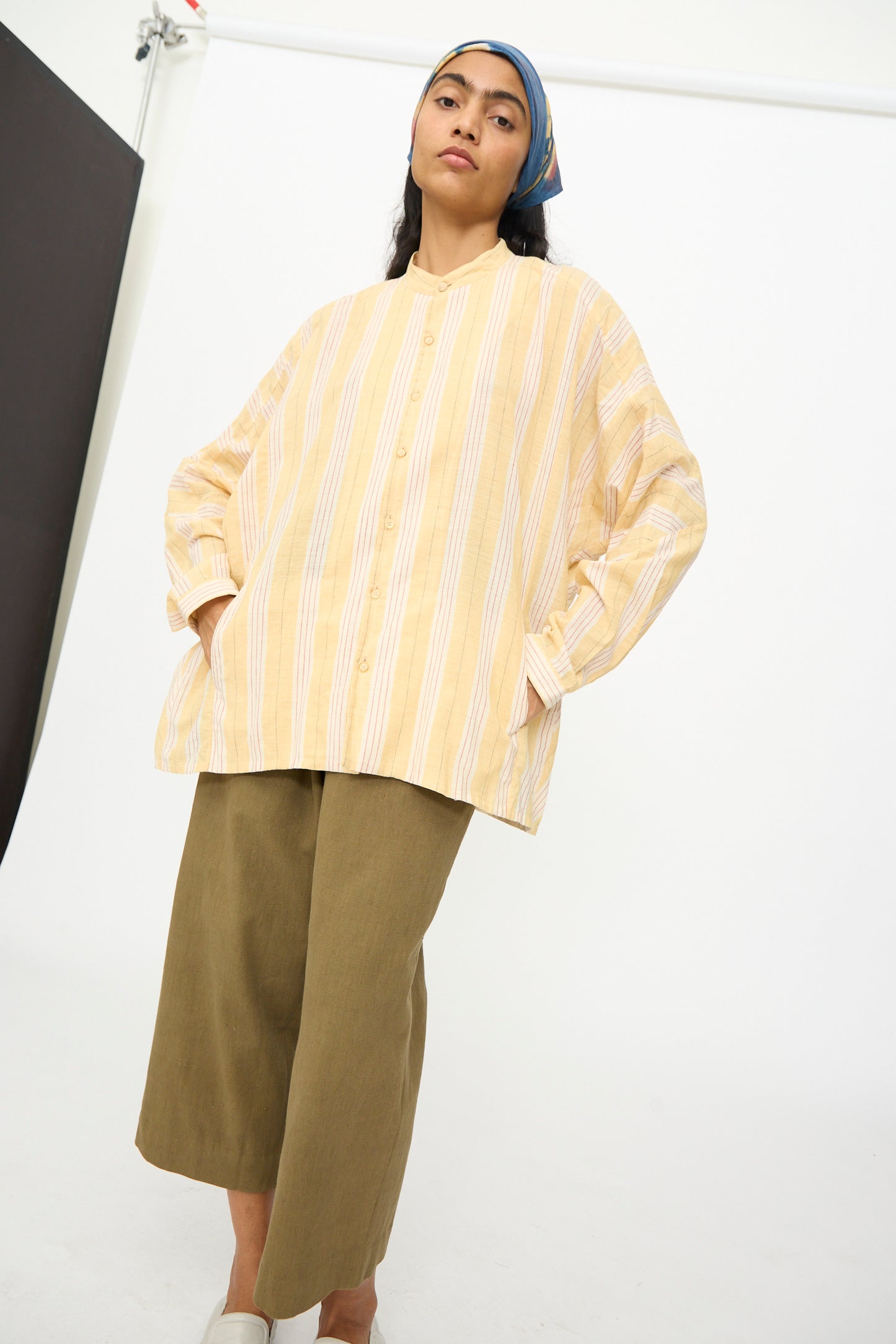 A person stands with hands in pockets, wearing the 11.11 Cotton Stripe Shirt in Mango Yellow, olive wide-leg pants, light shoes, and a blue headscarf against a plain studio background.