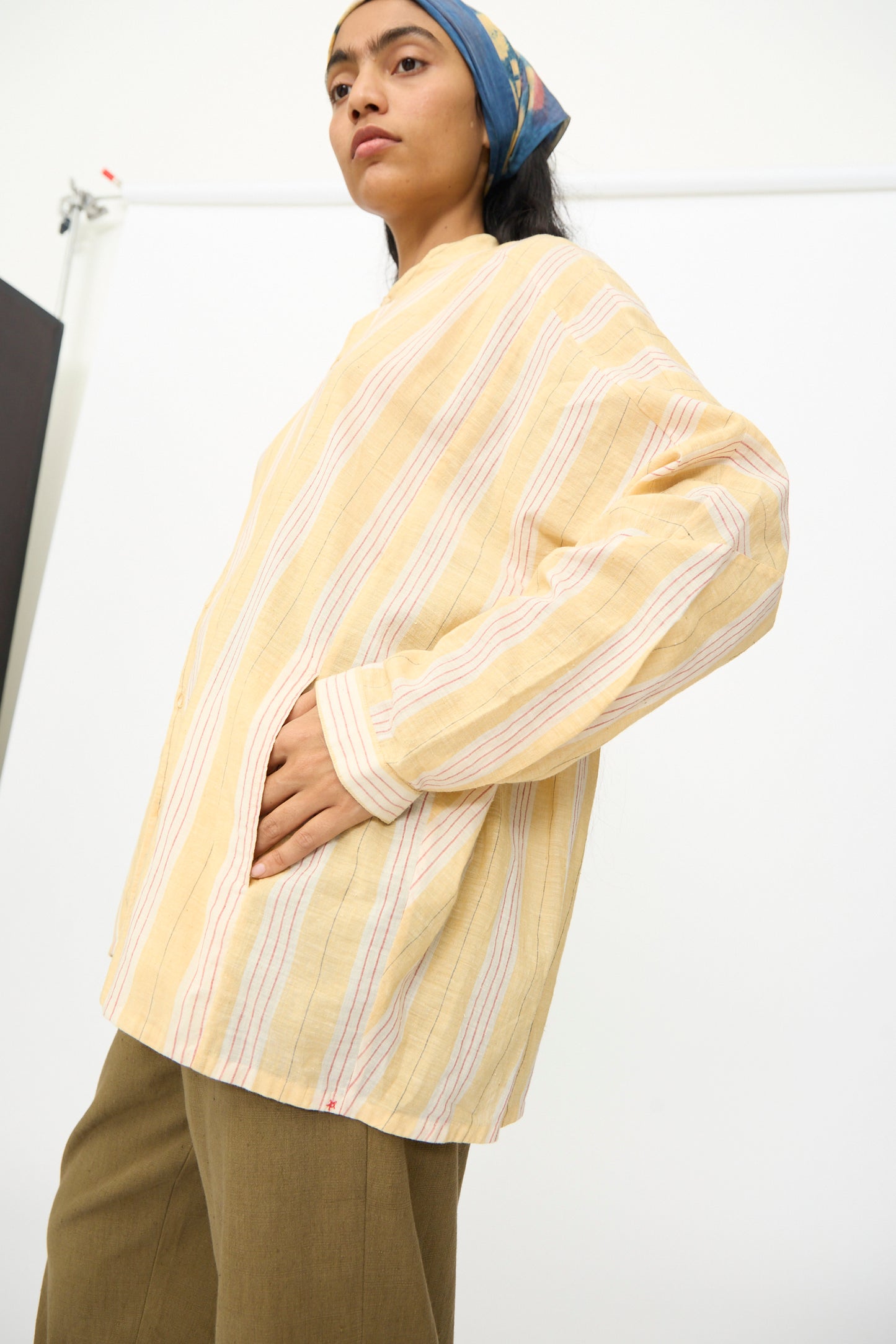 A person wearing the 11.11 Cotton Stripe Shirt in Mango Yellow with brown pants and a blue headscarf stands with one hand in their pocket against a plain background.