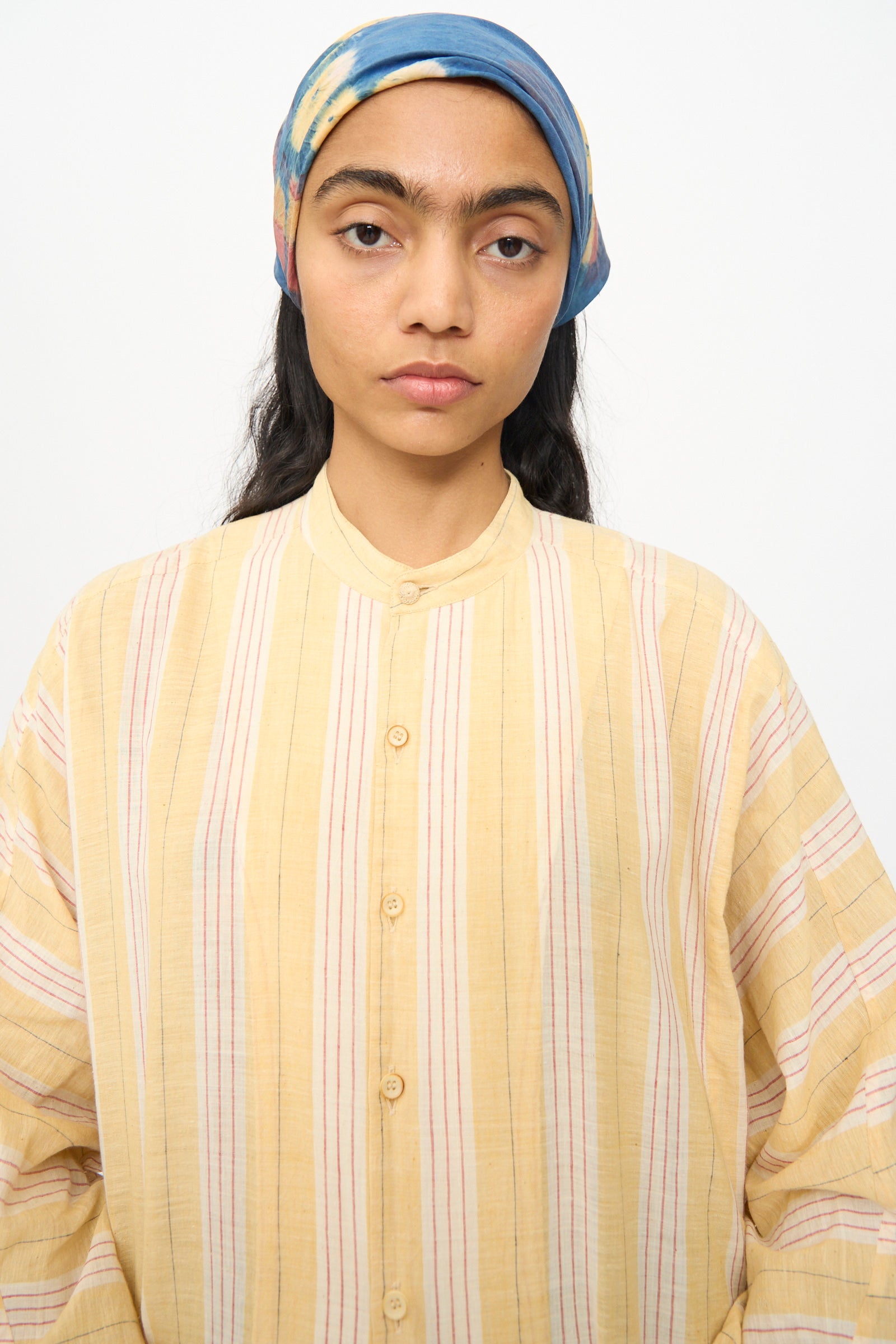 A person with long dark hair, wearing a blue scarf and the 11.11 Cotton Stripe Shirt in Mango Yellow, stands facing the camera against a plain background.