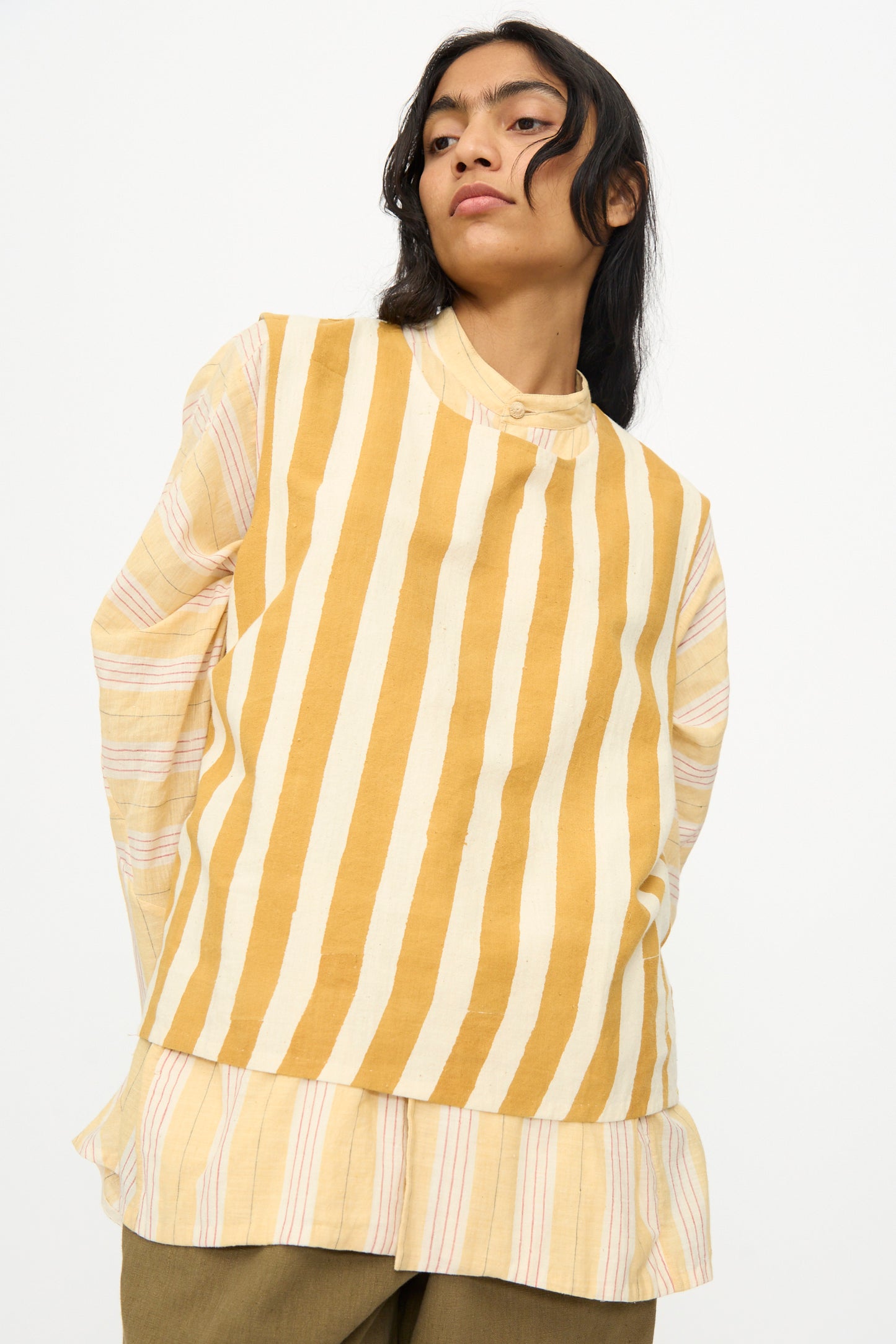 A person wearing the 11.11 Cotton Stripe Top in Mustard Yellow, featuring white vertical stripes and a cropped fit, layered over a long-sleeved shirt with pale pink and yellow stripes, stands against a plain background.