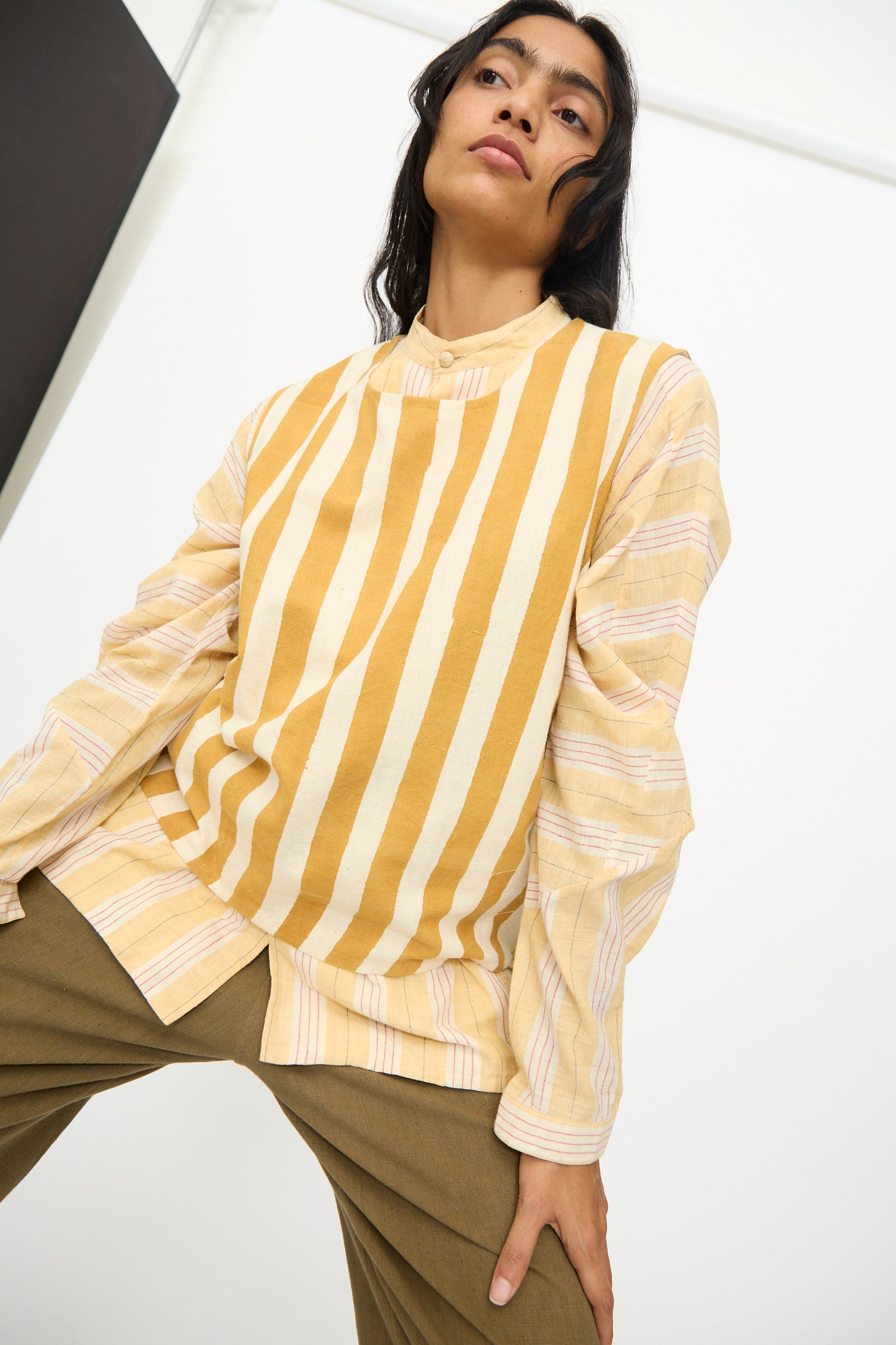 A person poses indoors in the 11.11 Cotton Stripe Top in Mustard Yellow—an oversized, handwoven tank with yellow and cream stripes—layered over a thinly striped long-sleeve shirt and paired with olive green pants.