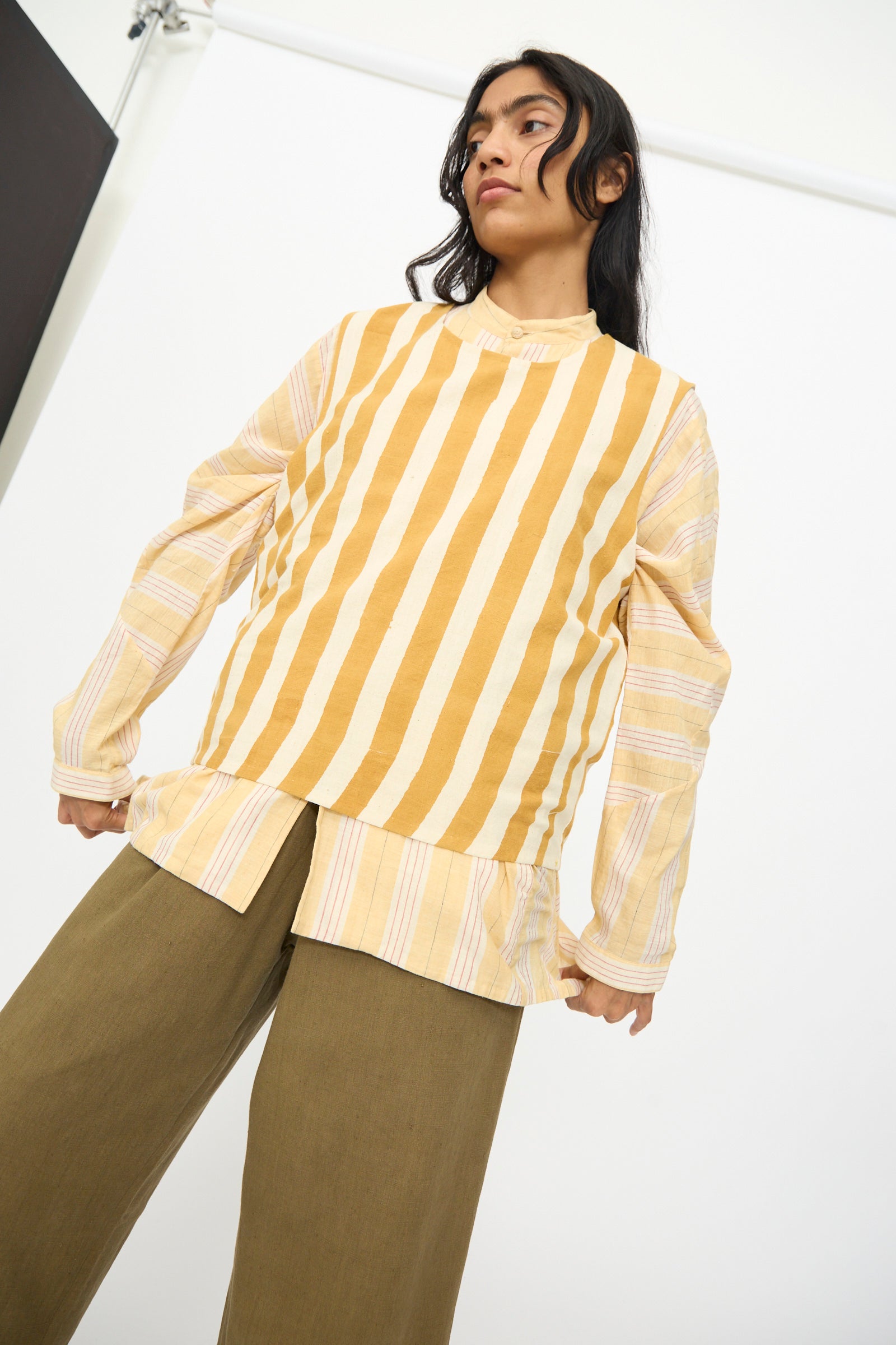 Person standing against a plain background, wearing the 11.11 Cotton Stripe Top in Mustard Yellow over a mustard tank top, paired with olive green wide-leg pants.