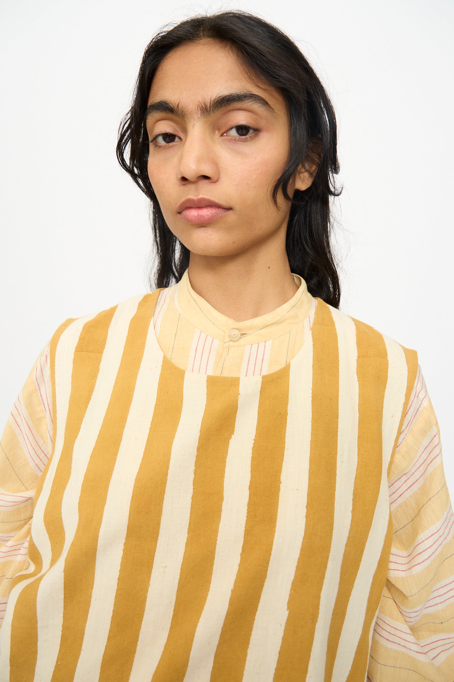 A person with long dark hair wears the 11.11 Cotton Stripe Top in Mustard Yellow, featuring white stripes and a mandarin collar, looking at the camera against a plain background.