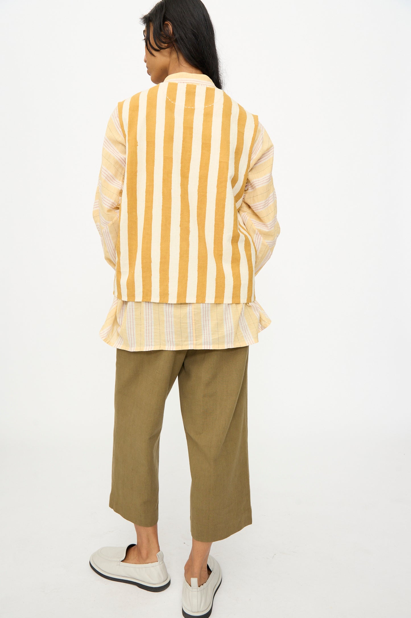 Model stands with back turned, wearing 11.11's Cotton Stripe Top in Mustard Yellow, a light layered shirt, olive green cropped pants, and white slip-on shoes against a plain background.