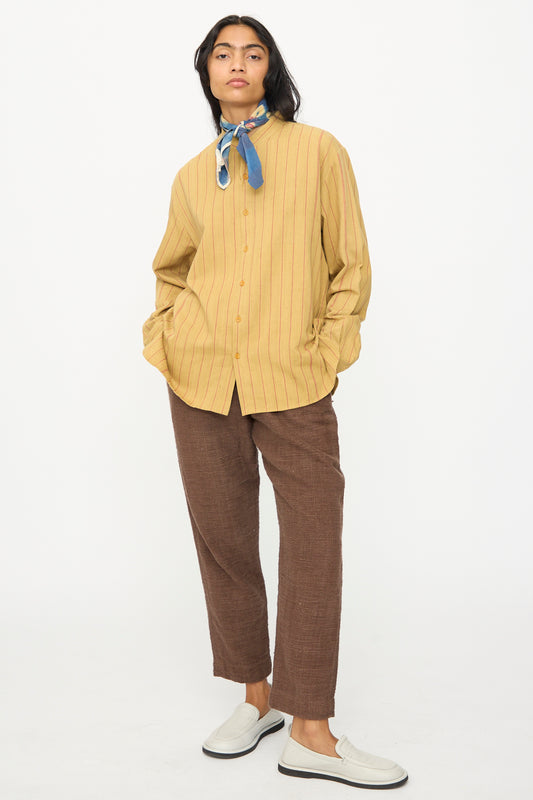 A person wears the 11.11 Lovers Shirt in Ochre with Red Stripe, brown pants, white slip-on shoes, and a blue neck scarf, standing with hands in pockets against a plain white background.