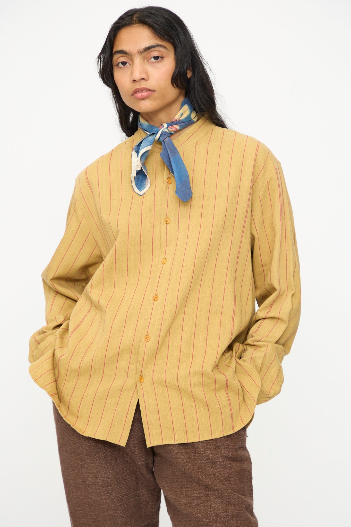 Model wears the Lovers Shirt in Ochre with Red Stripe by 11.11, paired with brown pants and a blue patterned scarf, standing against a plain background.
