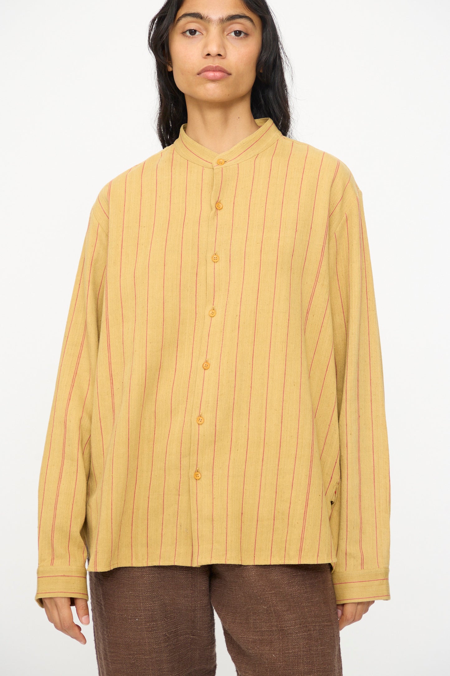 A person wears the 11.11 Lovers Shirt in Ochre with Red Stripe, a relaxed fit yellow button-up with red vertical stripes, paired with brown pants, standing against a plain white background.