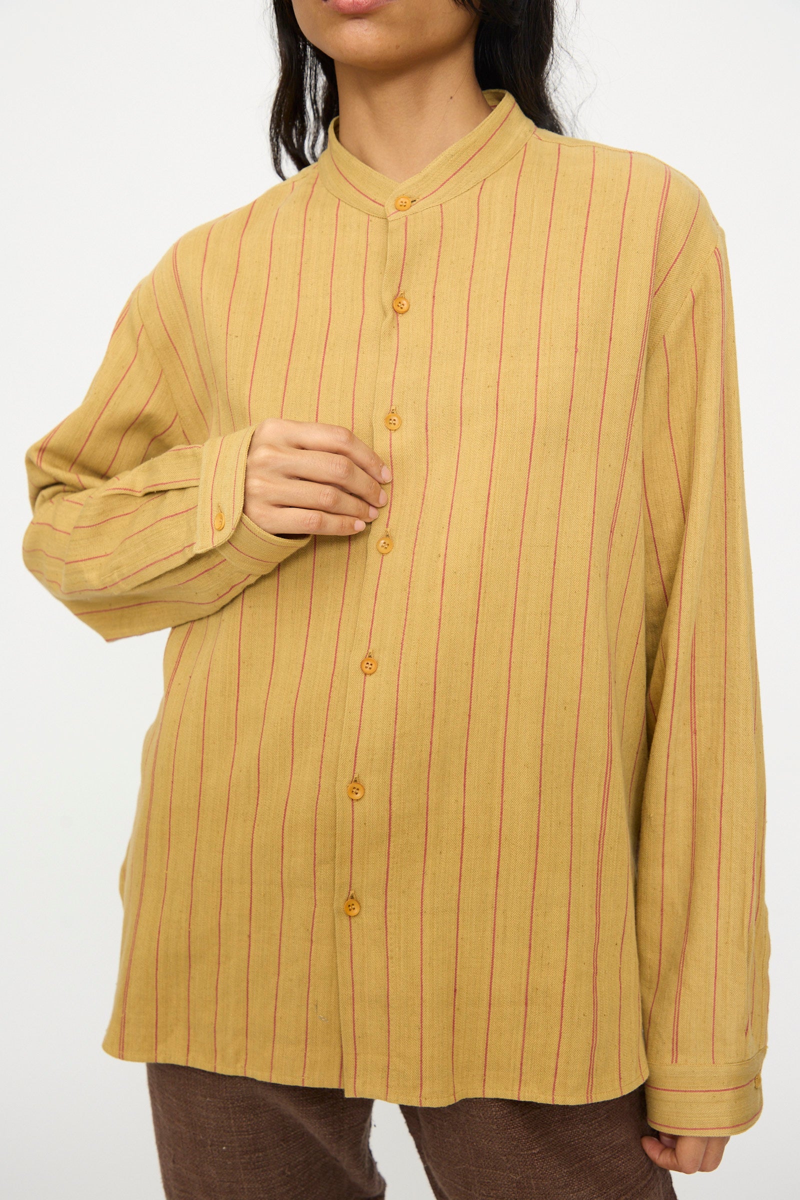 A person wears the 11.11 Lovers Shirt in Ochre with Red Stripe, a relaxed fit button up featuring brown buttons and a band collar, standing against a plain background.