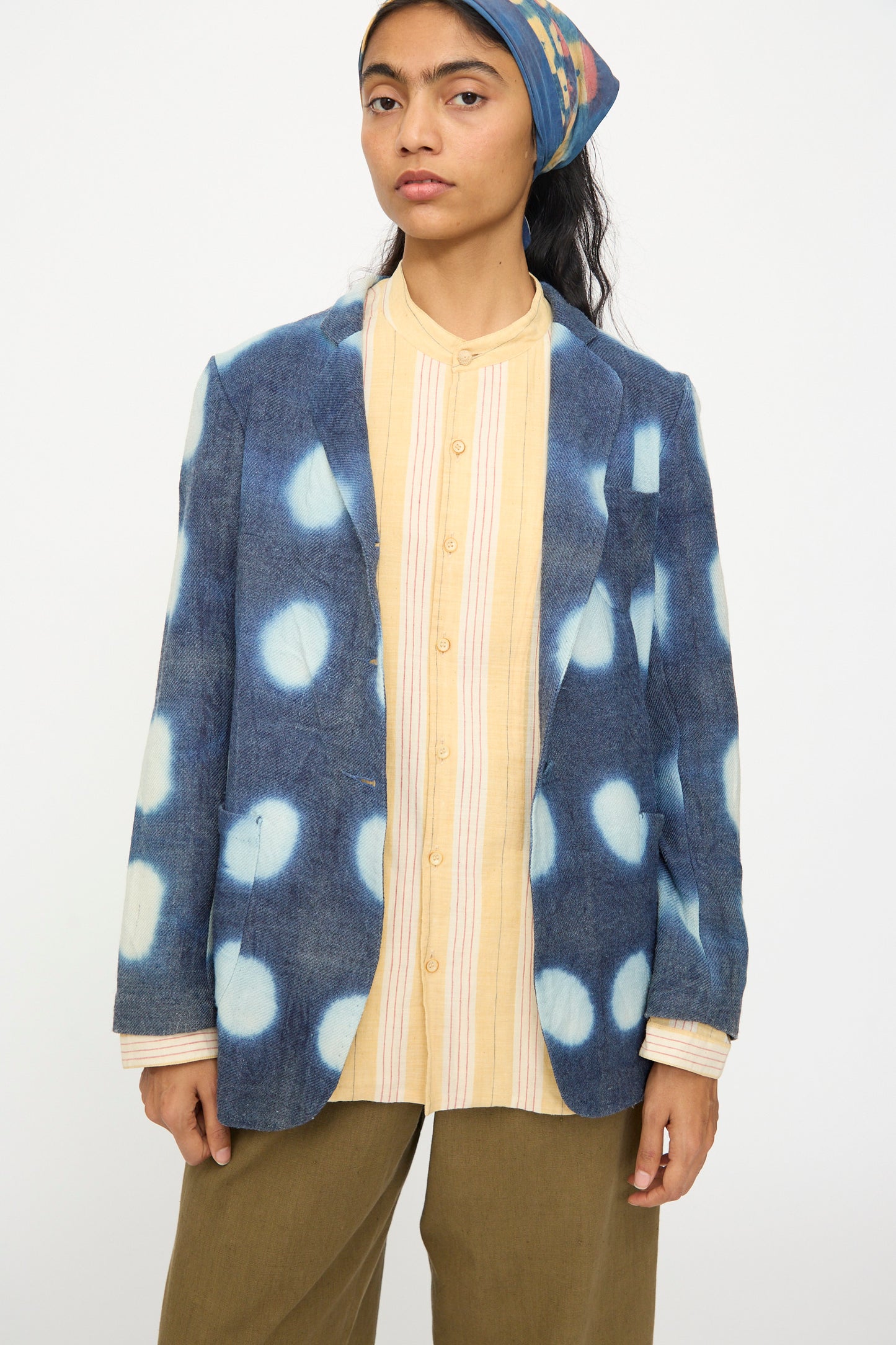 A woman wearing the 11.11 Nico Clamp Dye Merino Wool Jacket in Indigo, a yellow-striped button-up shirt, olive pants, and a patterned headscarf stands against a plain background.