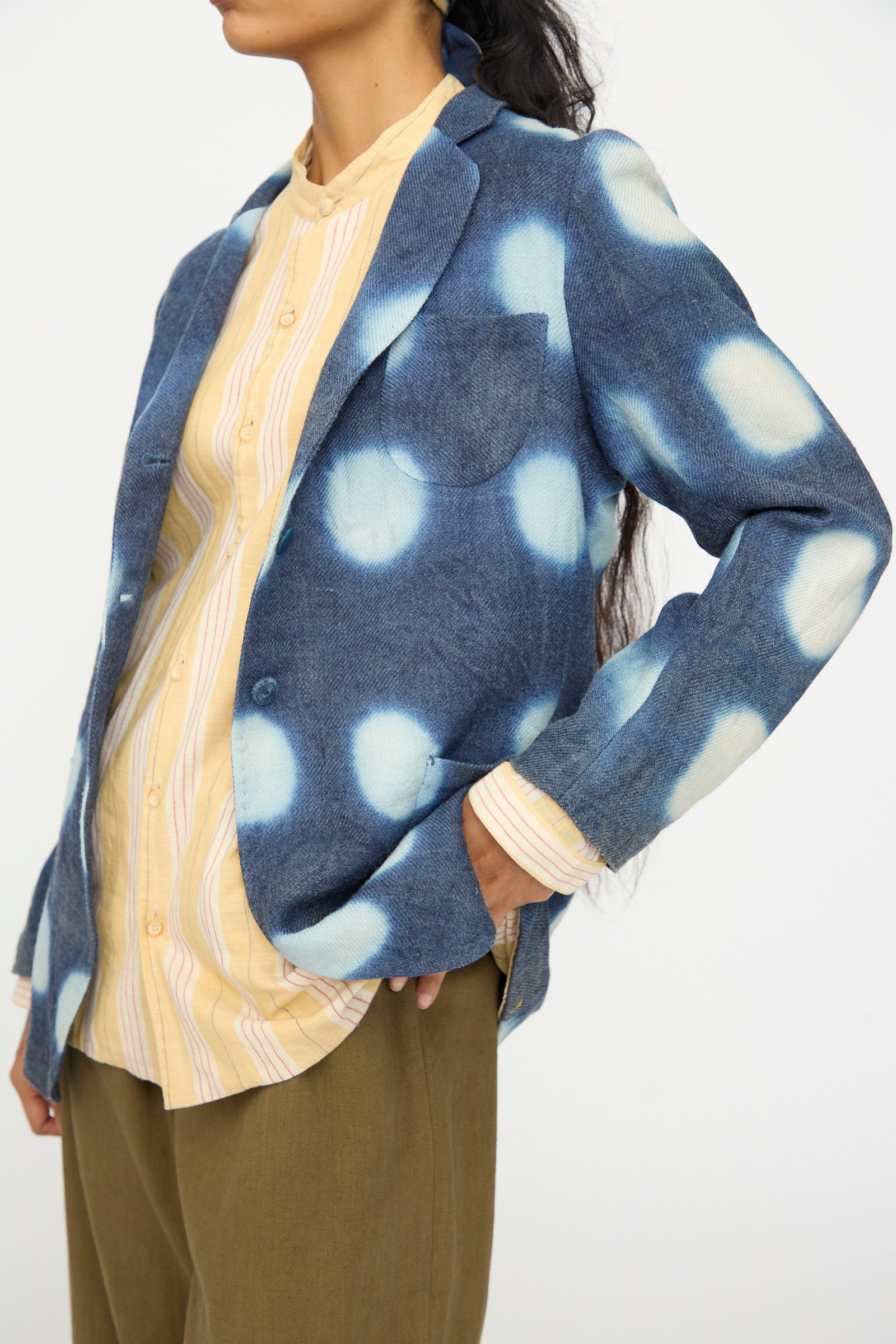 A person wears the 11.11 Nico Clamp Dye Merino Wool Jacket in Indigo, featuring white circular patterns, paired with a light yellow striped shirt and olive green pants against a plain background.