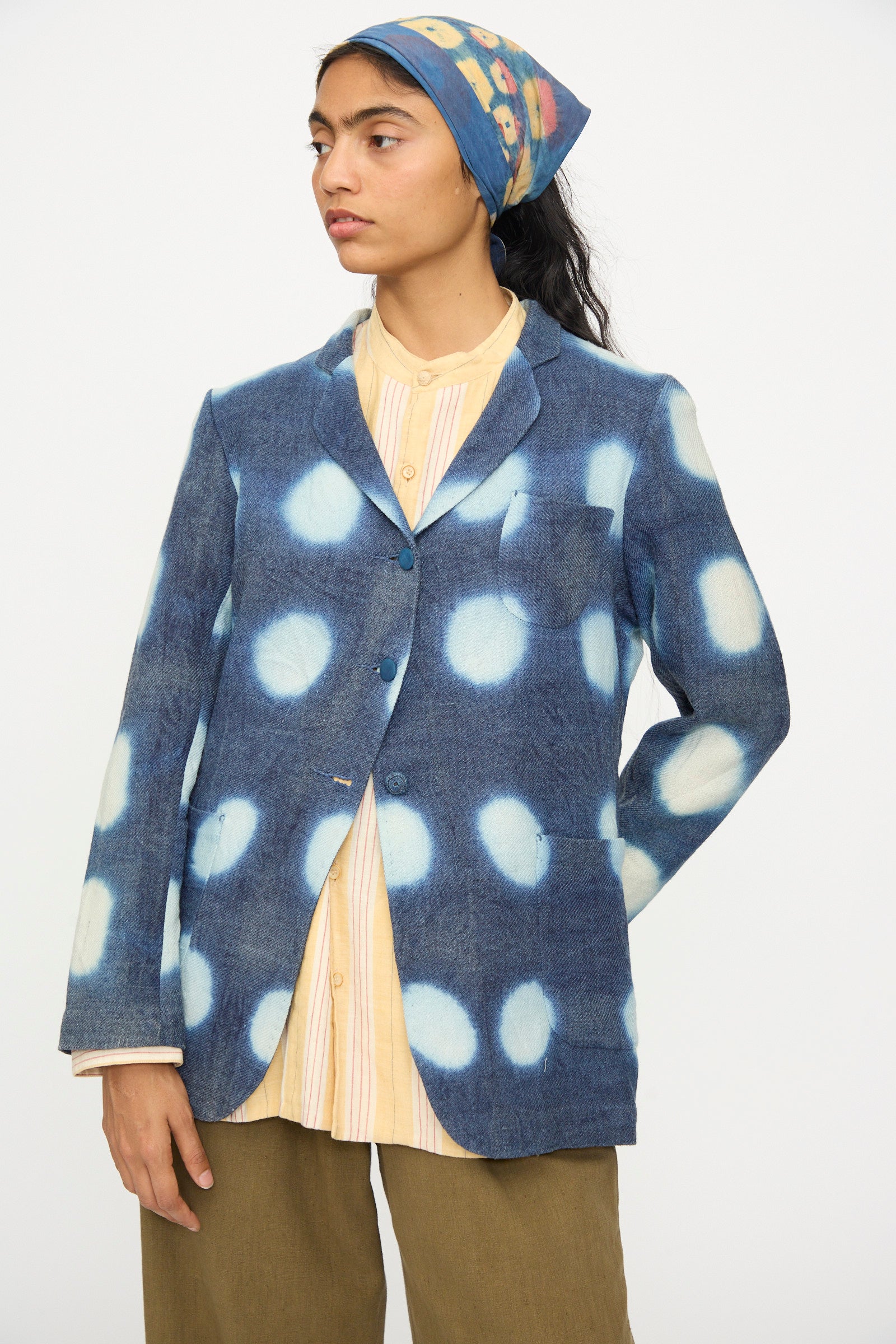 A person wearing the 11.11 Nico Clamp Dye Merino Wool Jacket in Indigo, paired with a yellow striped shirt, olive green pants, and a blue headscarf with yellow designs, stands against a plain background.
