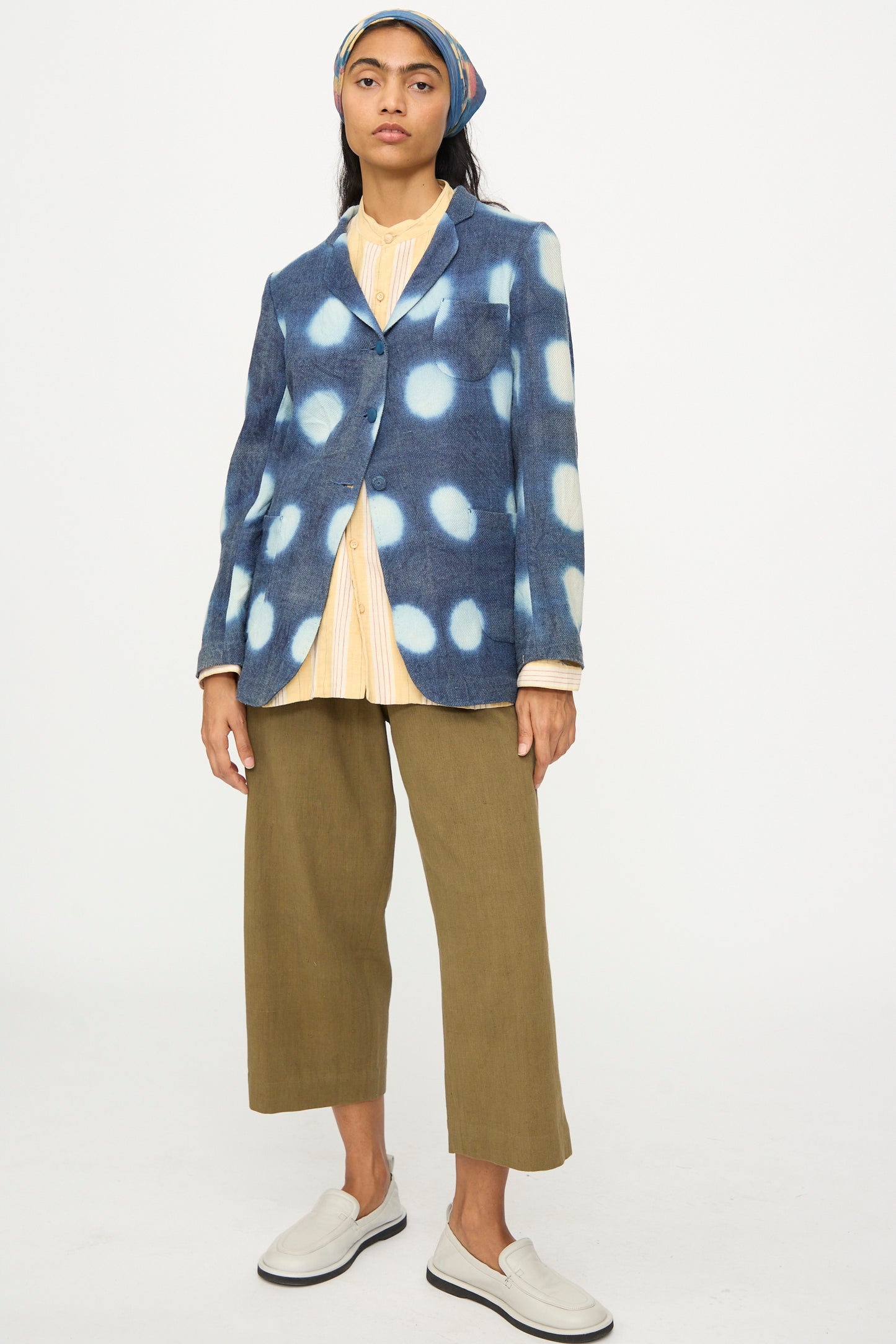 A person wears the 11.11 Nico Clamp Dye Merino Wool Jacket in Indigo, paired with a yellow striped shirt, olive green wide-leg pants, white slip-on shoes, and a headscarf against a plain white background.