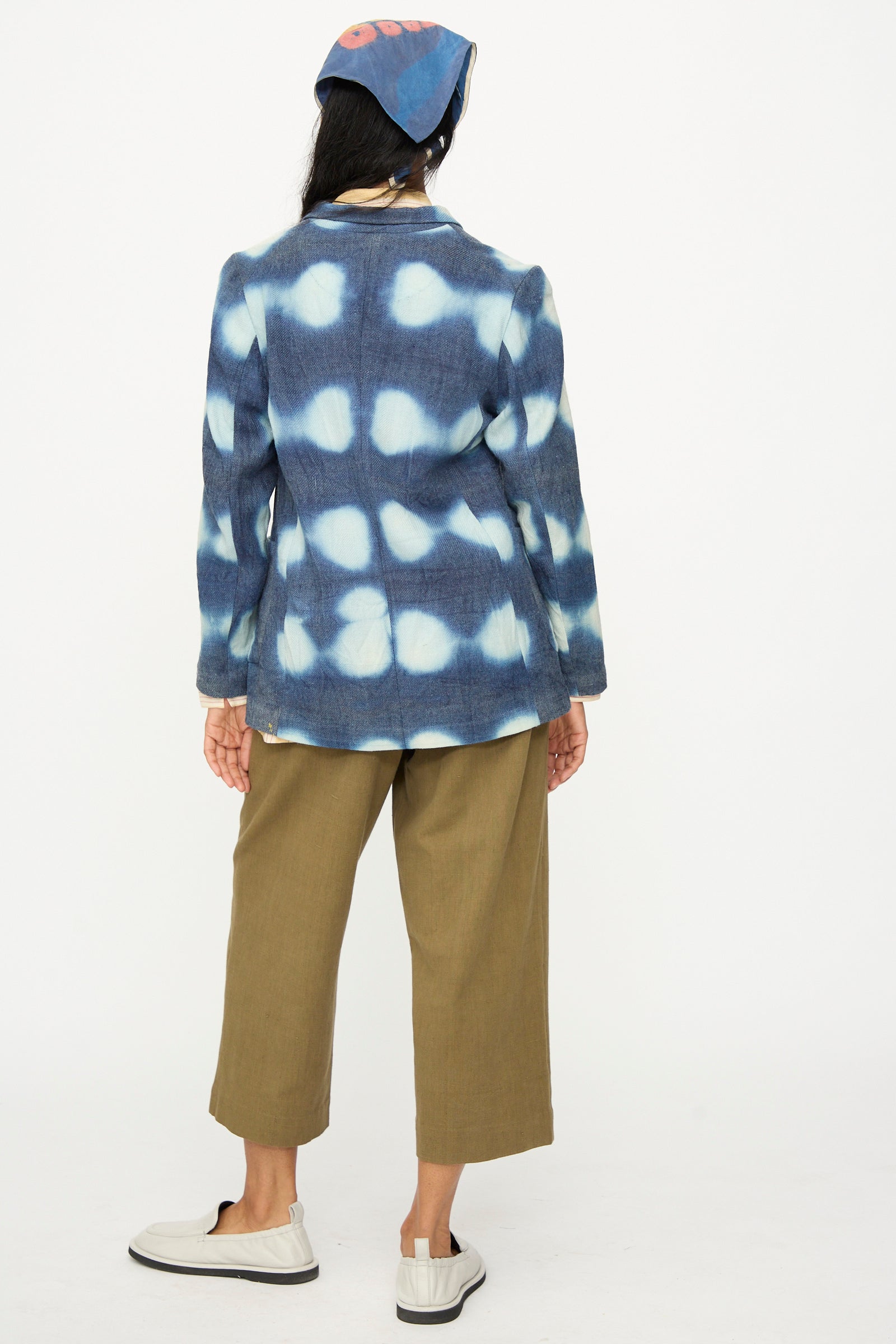 A person stands facing away, wearing the 11.11 Nico Clamp Dye Merino Wool Jacket in Indigo, brown wide-leg pants, white shoes, and a blue headscarf with a pink design.