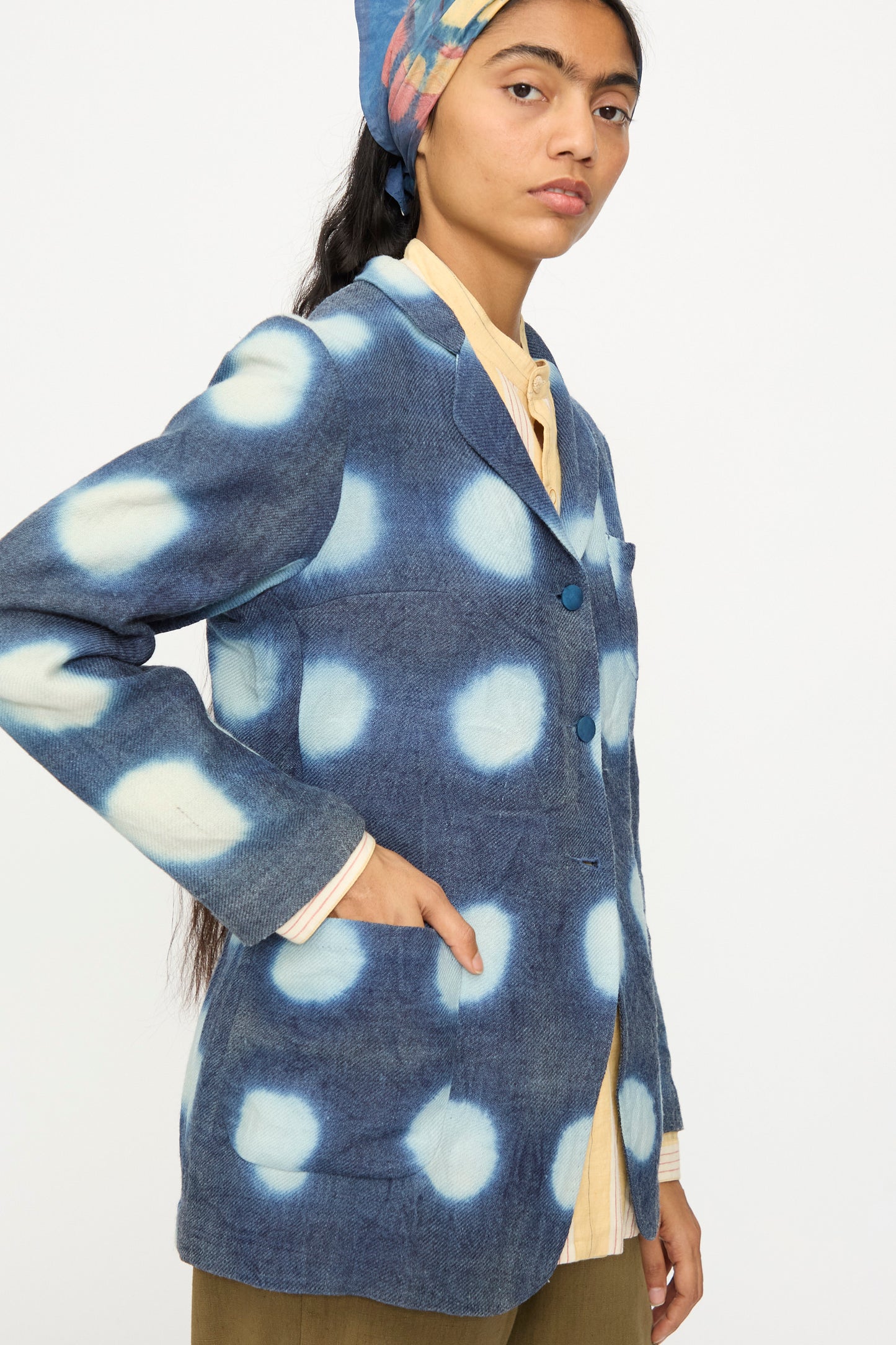 A person wearing the 11.11 Nico Clamp Dye Merino Wool Jacket in Indigo, styled with a yellow shirt and a multicolored headscarf, stands against a plain background.