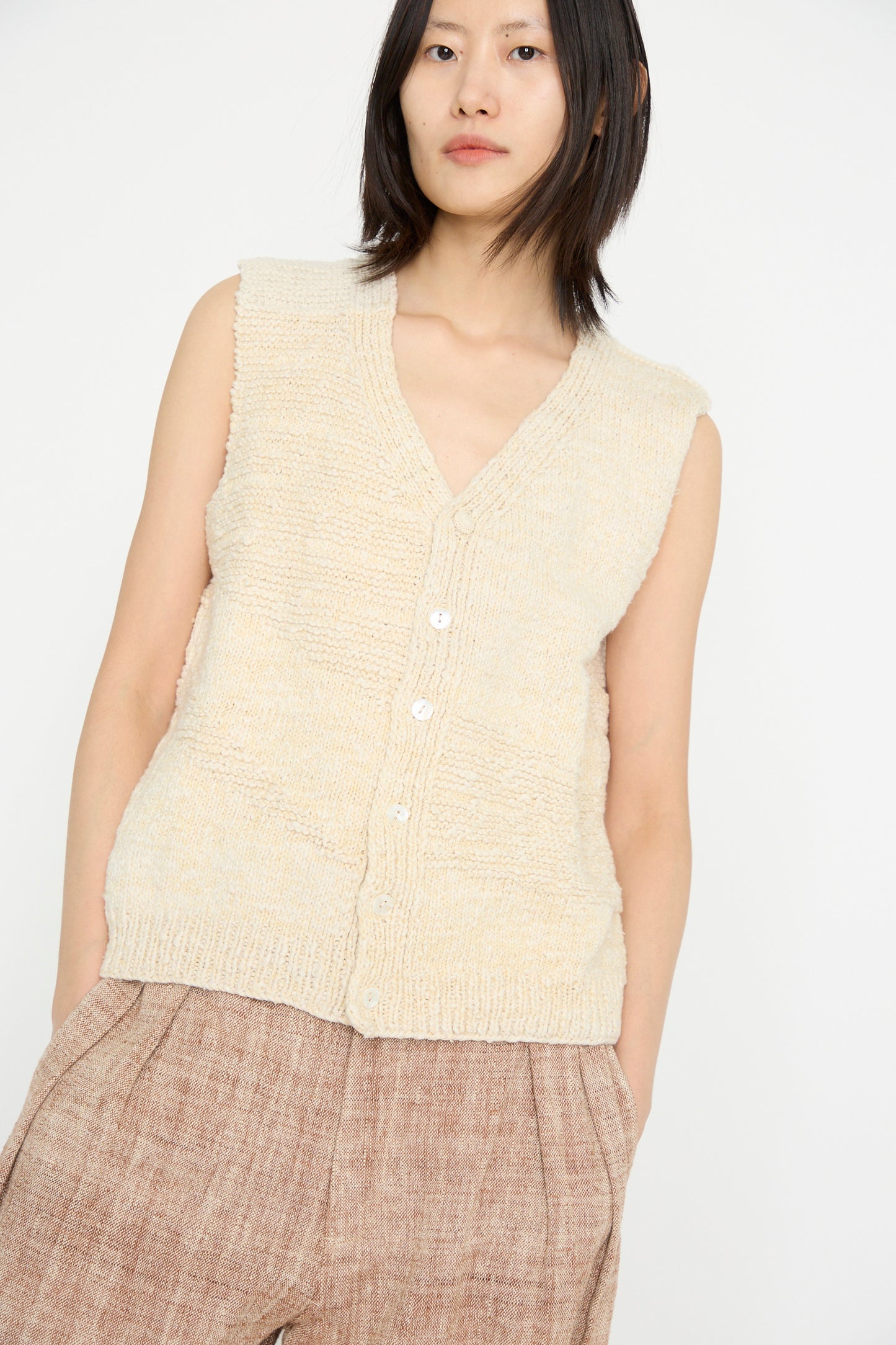 A person stands against a plain white background, wearing the Oro Vest in Ecru by 11.11, made of organic cotton and paired with brown trousers.