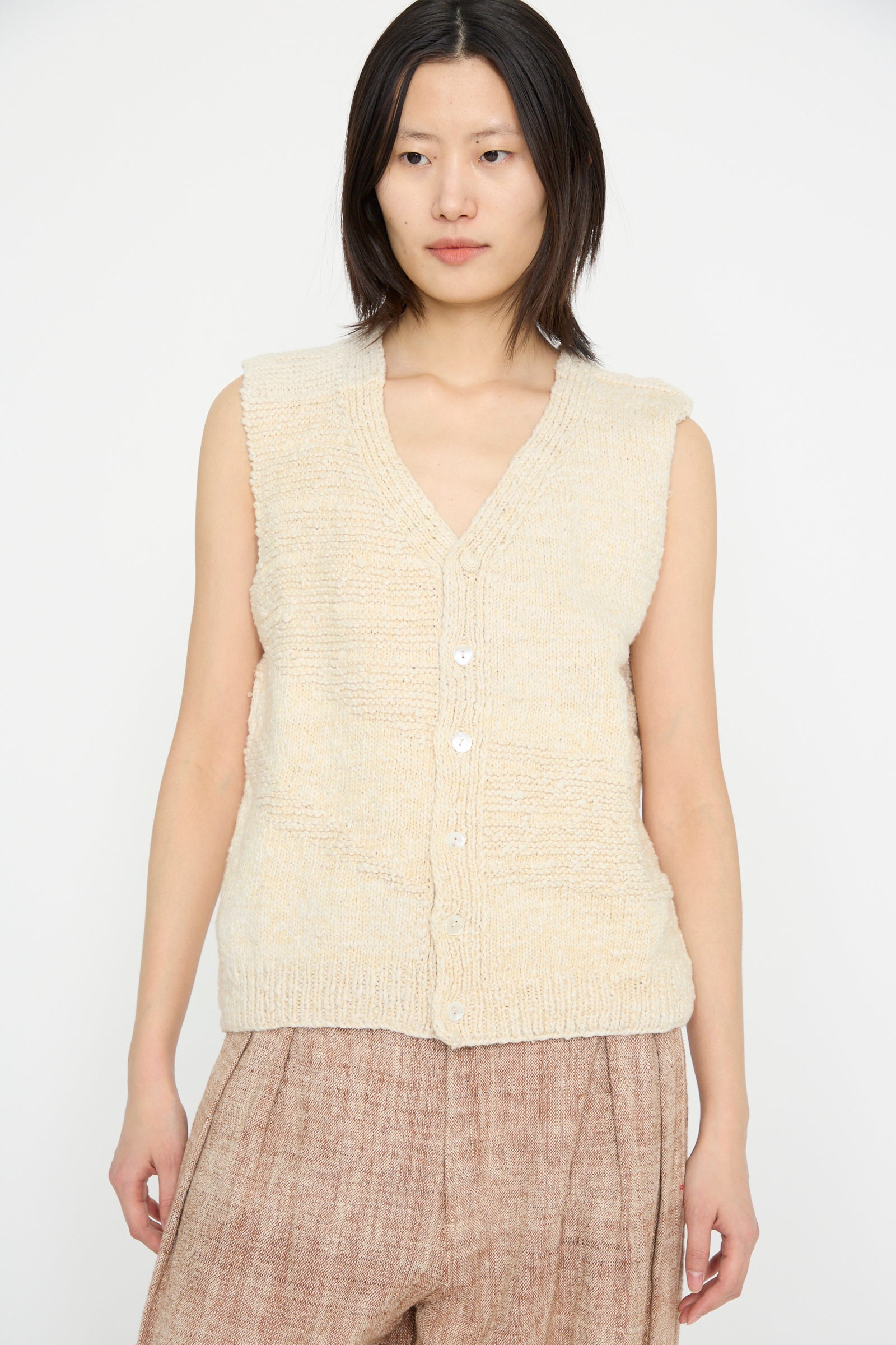 A person is wearing the Oro Vest in Ecru by 11.11, a beige sleeveless knit top made of luxurious Merino wool, paired with brown pants against a plain white background.