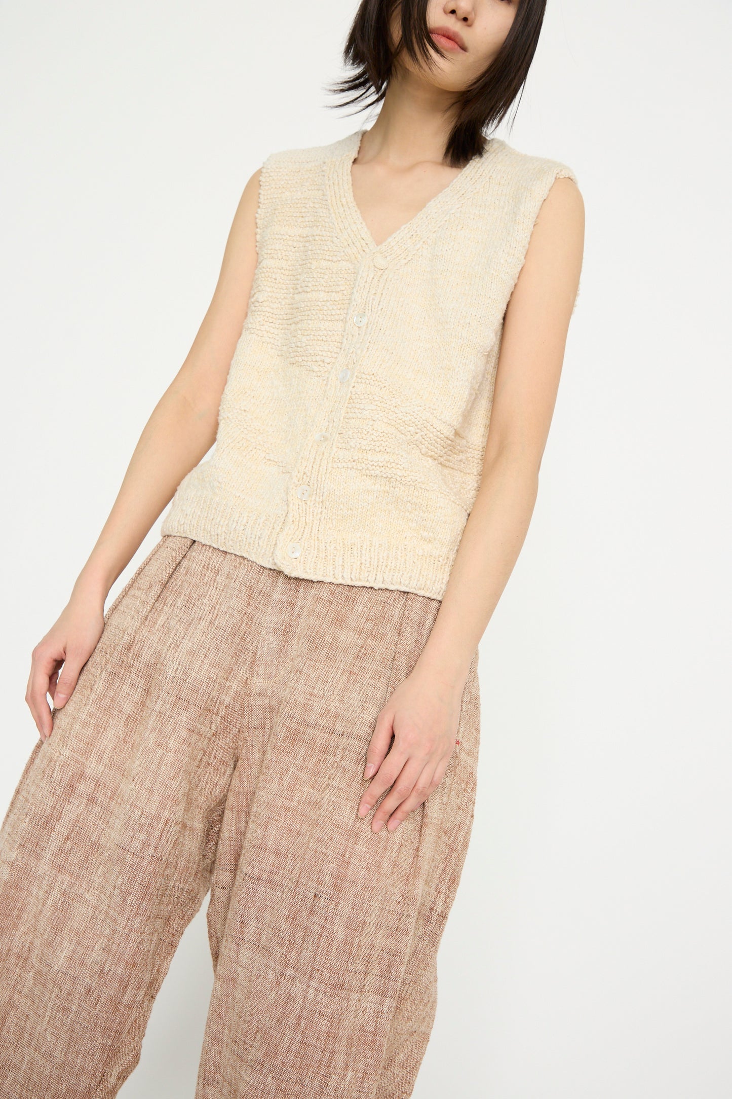 A person poses against a plain background, wearing the Oro Vest in Ecru by 11.11 and brown wide-leg pants.