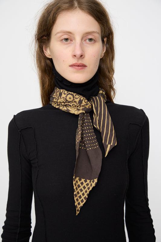 A woman in a black long-sleeve top wears the 11.11 Silk Bandana in Charcoal Block Print, tied around her neck, standing against a plain light background.